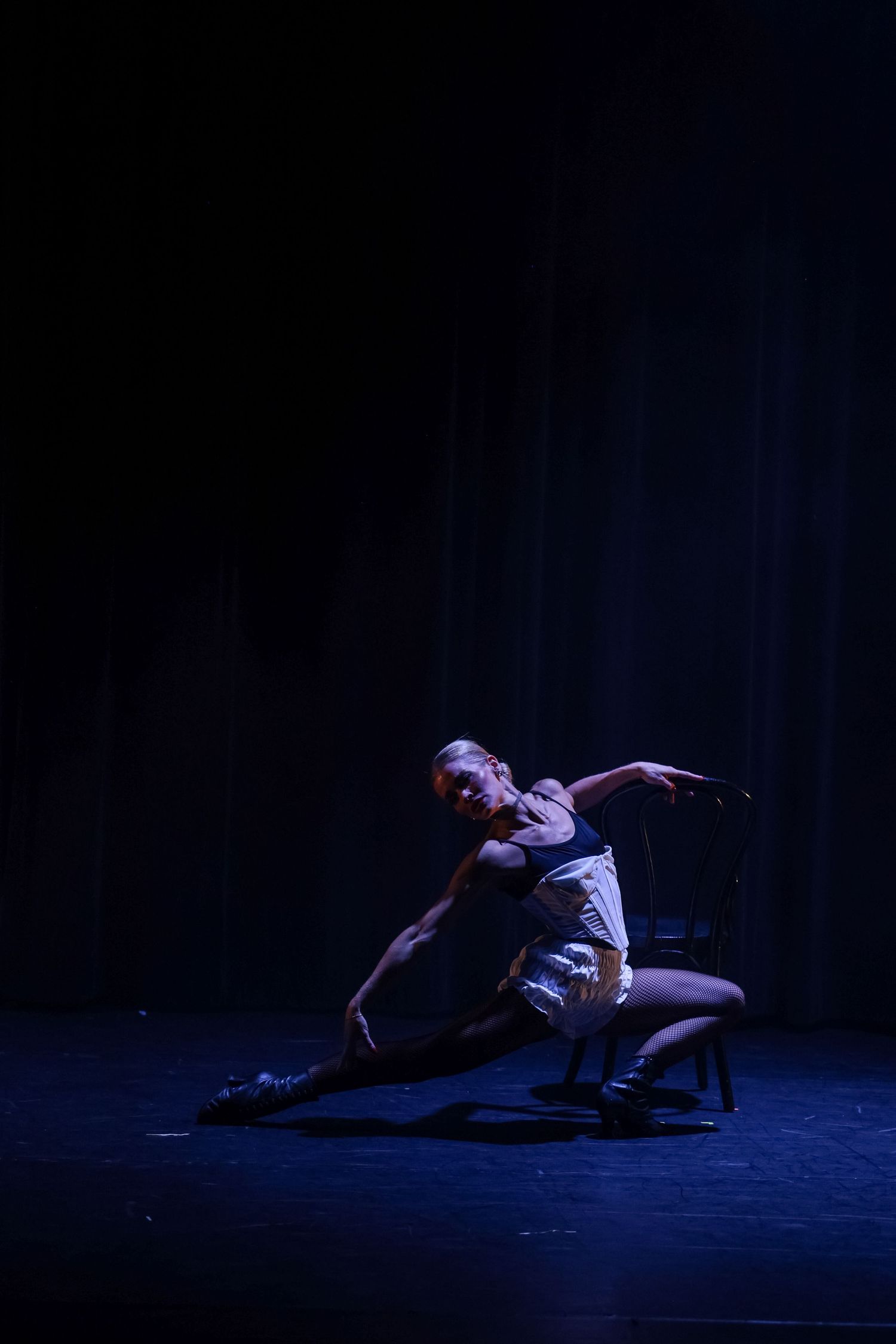 A dancer performs an expressive contemporary routine on a dark stage, elegantly posed beside a chair in white shorts.