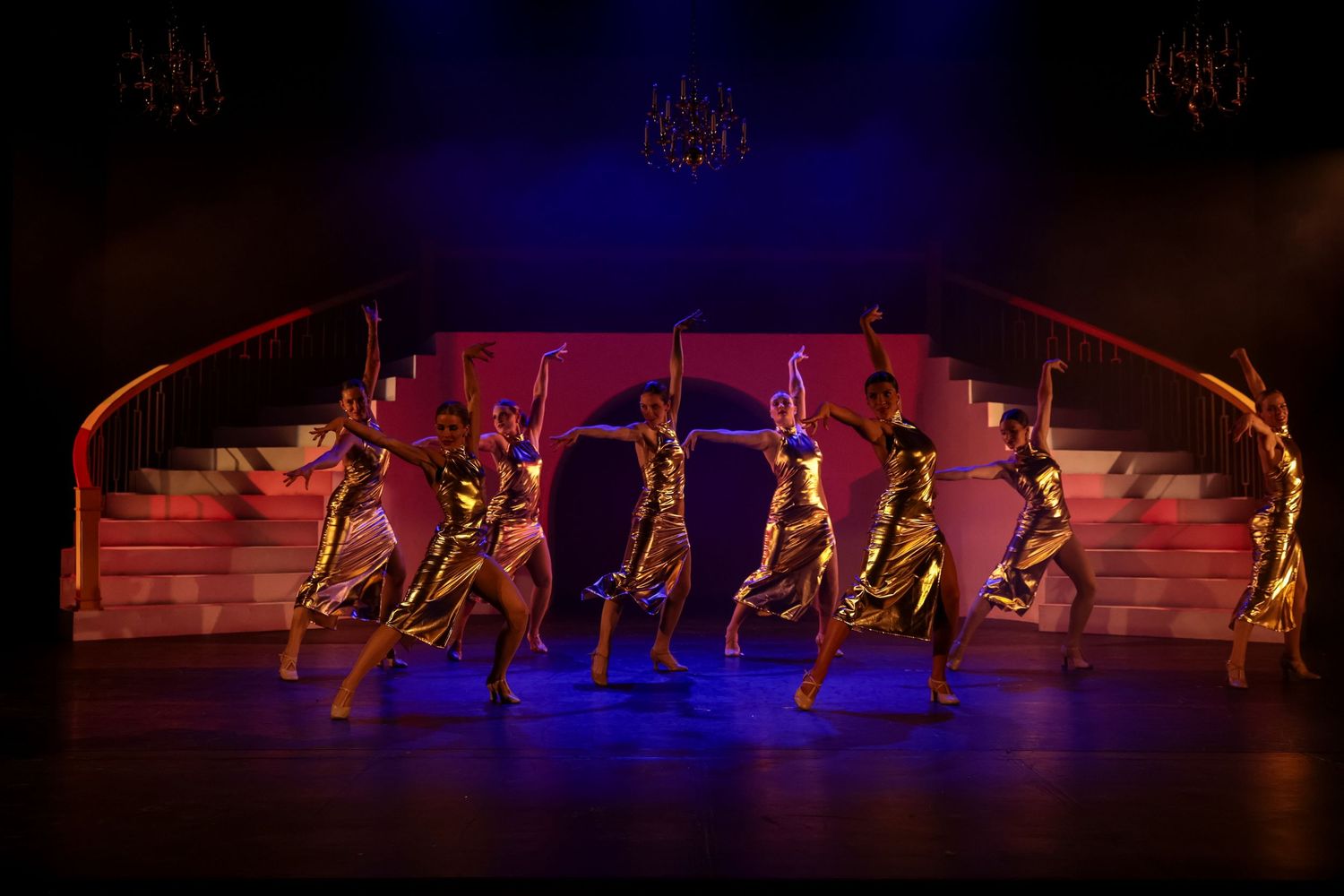 Seven dancers in gold dresses perform on a theatrical stage with pink backdrop and elegant staircase under dramatic lighting.