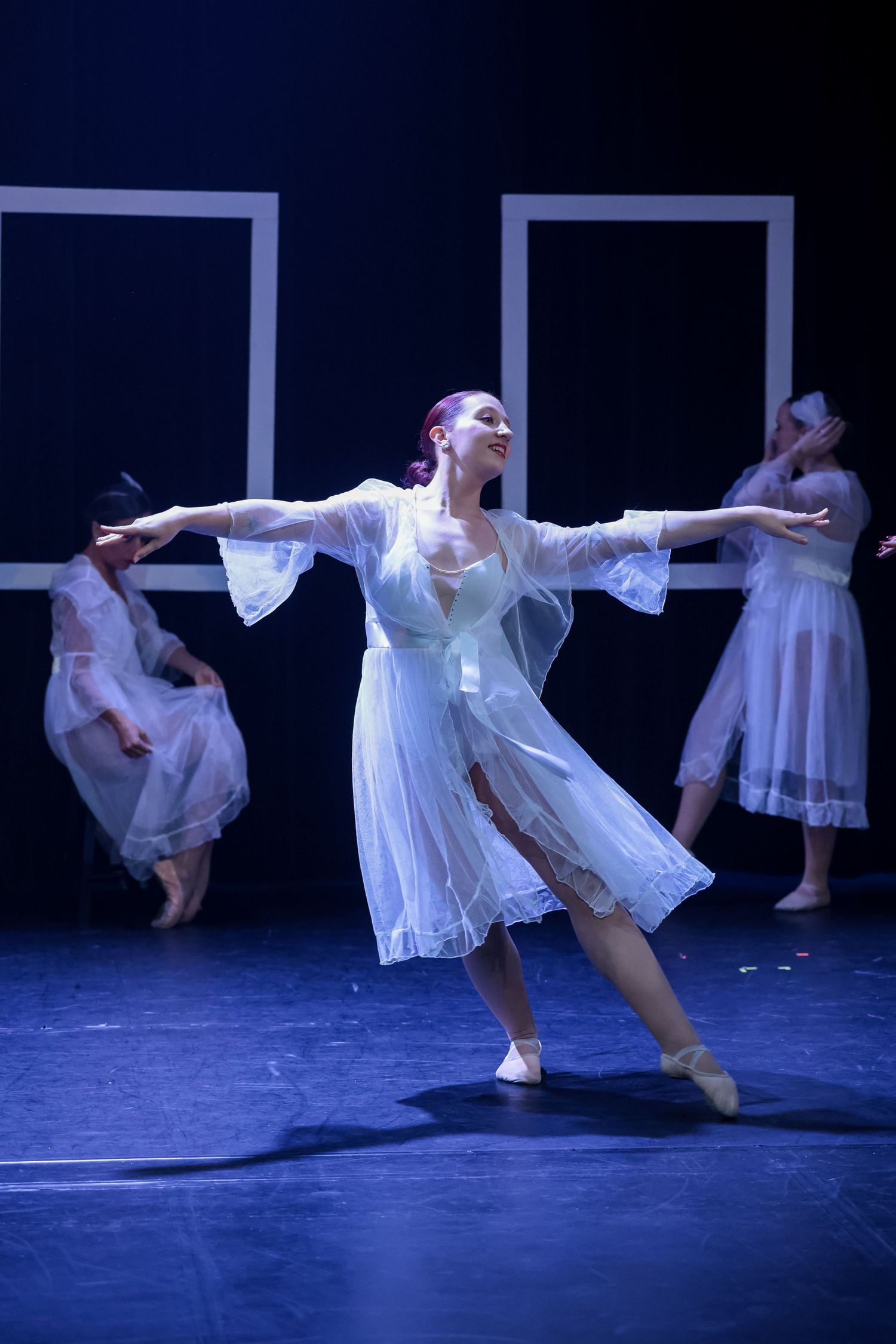 Graceful ballet dancer in white dress performs on stage with arms extended, blue lighting, and dancers in background.