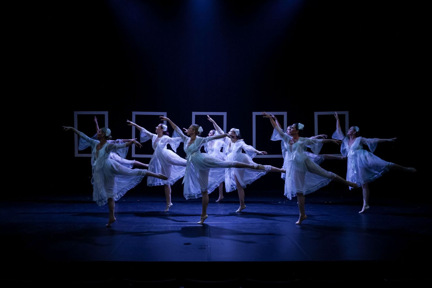 Ballet dancers in white dresses perform on a dark stage with illuminated square frames in the background.