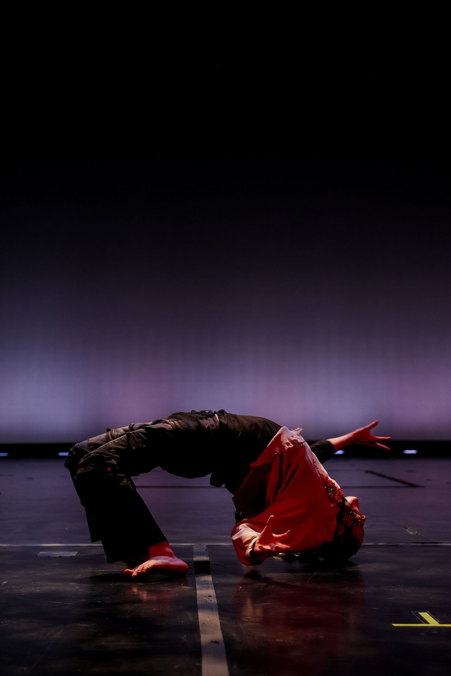 Breakdancer performing a backbend move on a dark stage, wearing red top and black pants under dramatic purple lighting.