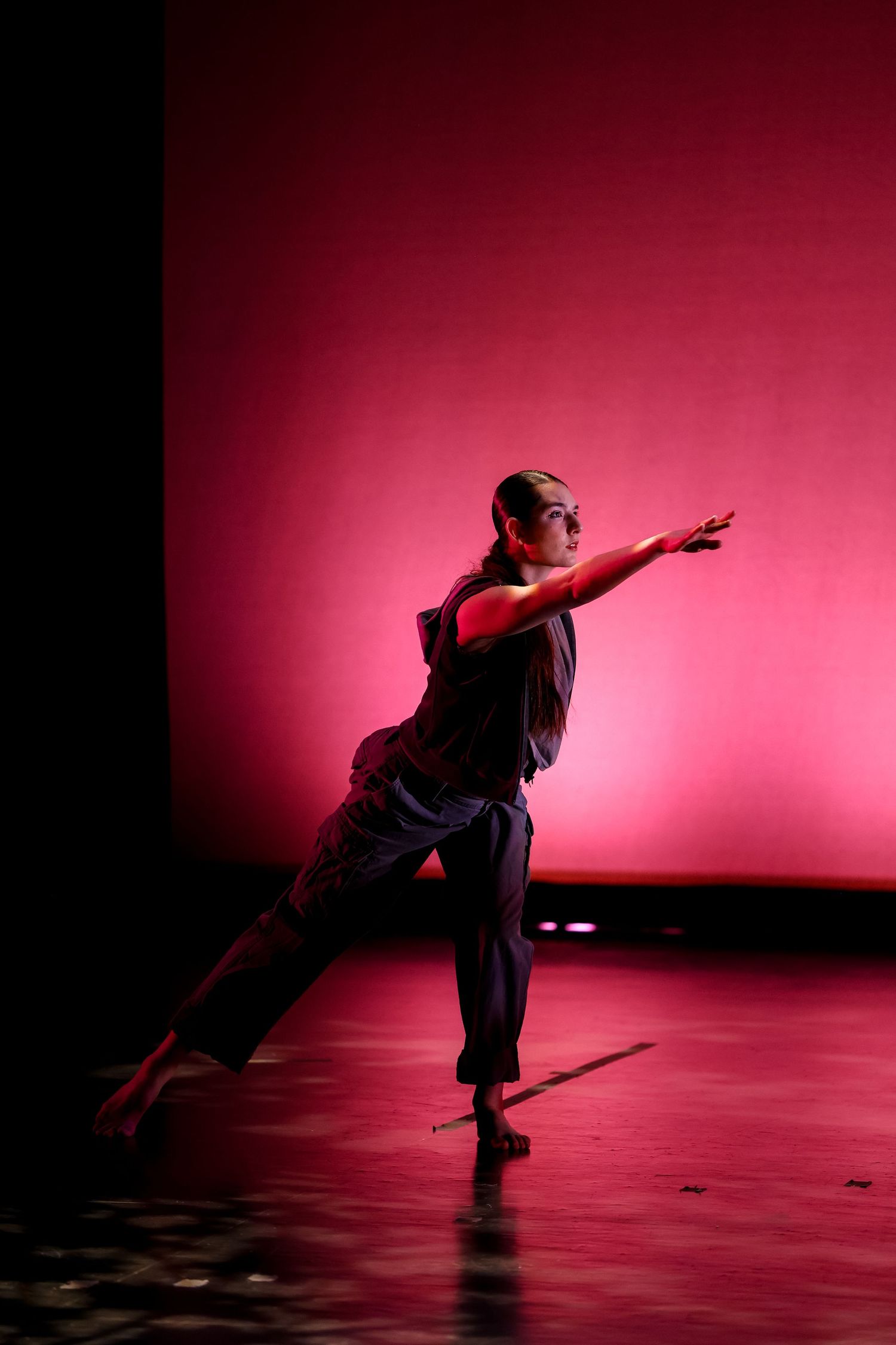 A dancer performs on a red-lit stage, extending arms gracefully in dark clothing during a modern dance performance.