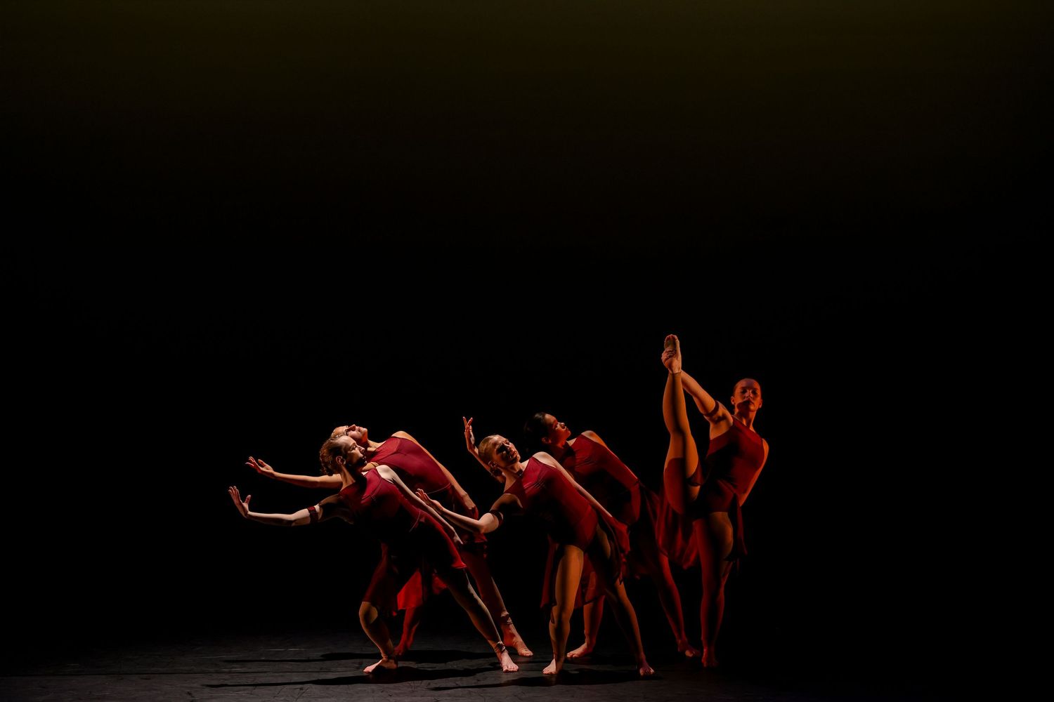 Dancers in red costumes perform a dramatic contemporary dance piece on a dark stage under warm spotlight.