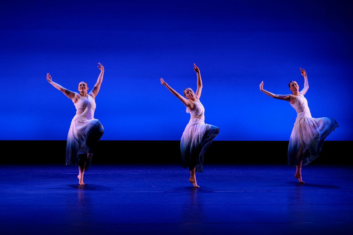 Three ballet dancers in white dresses perform gracefully on a blue-lit stage in perfect synchronized formation.