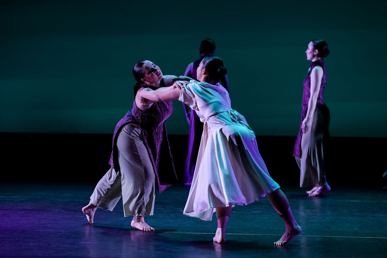 Dancers perform a contemporary dance piece on a dimly lit stage with teal and purple lighting.