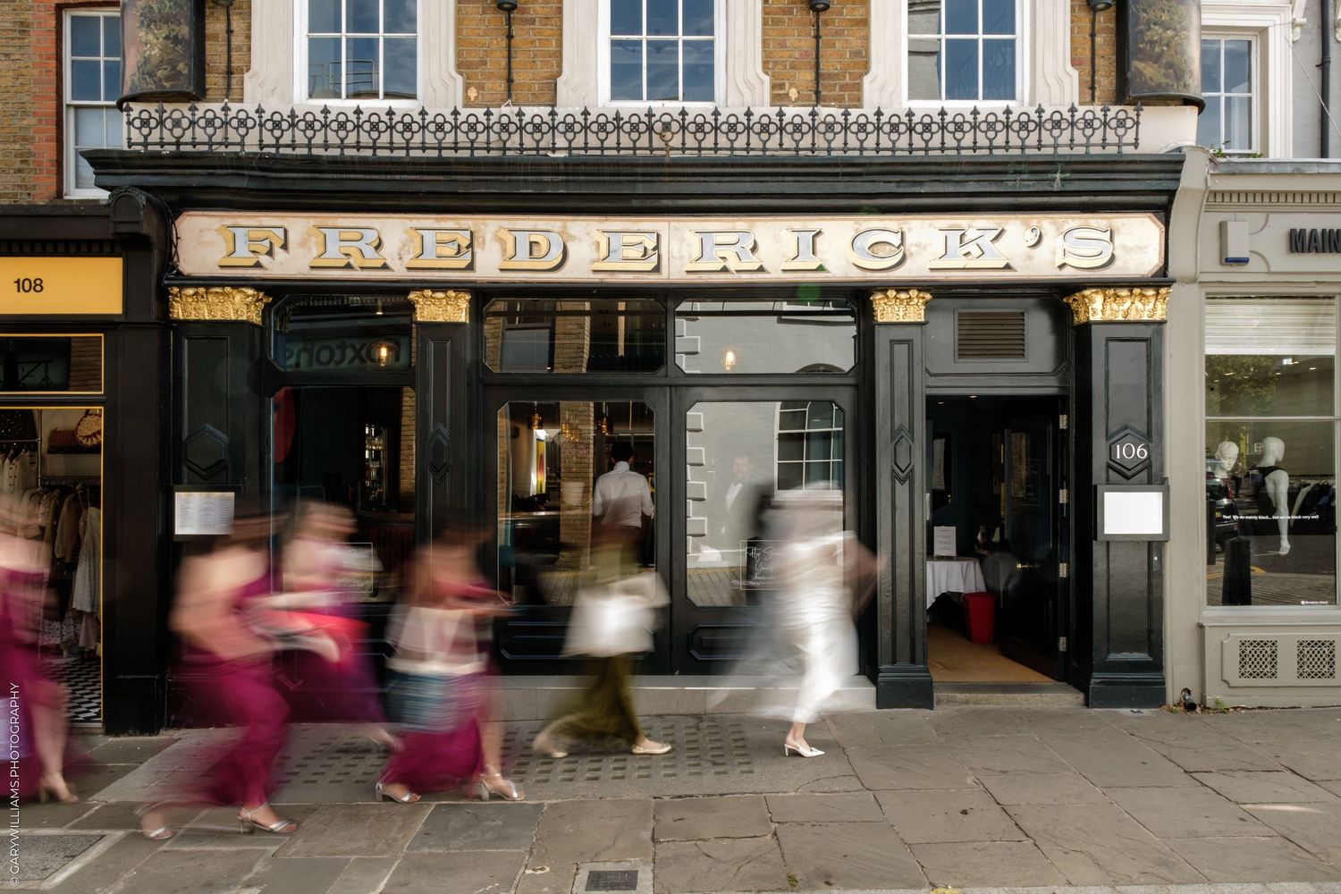 Blurred pedestrians walk past Frederick's restaurant shop front on a busy London street on a sunny day.