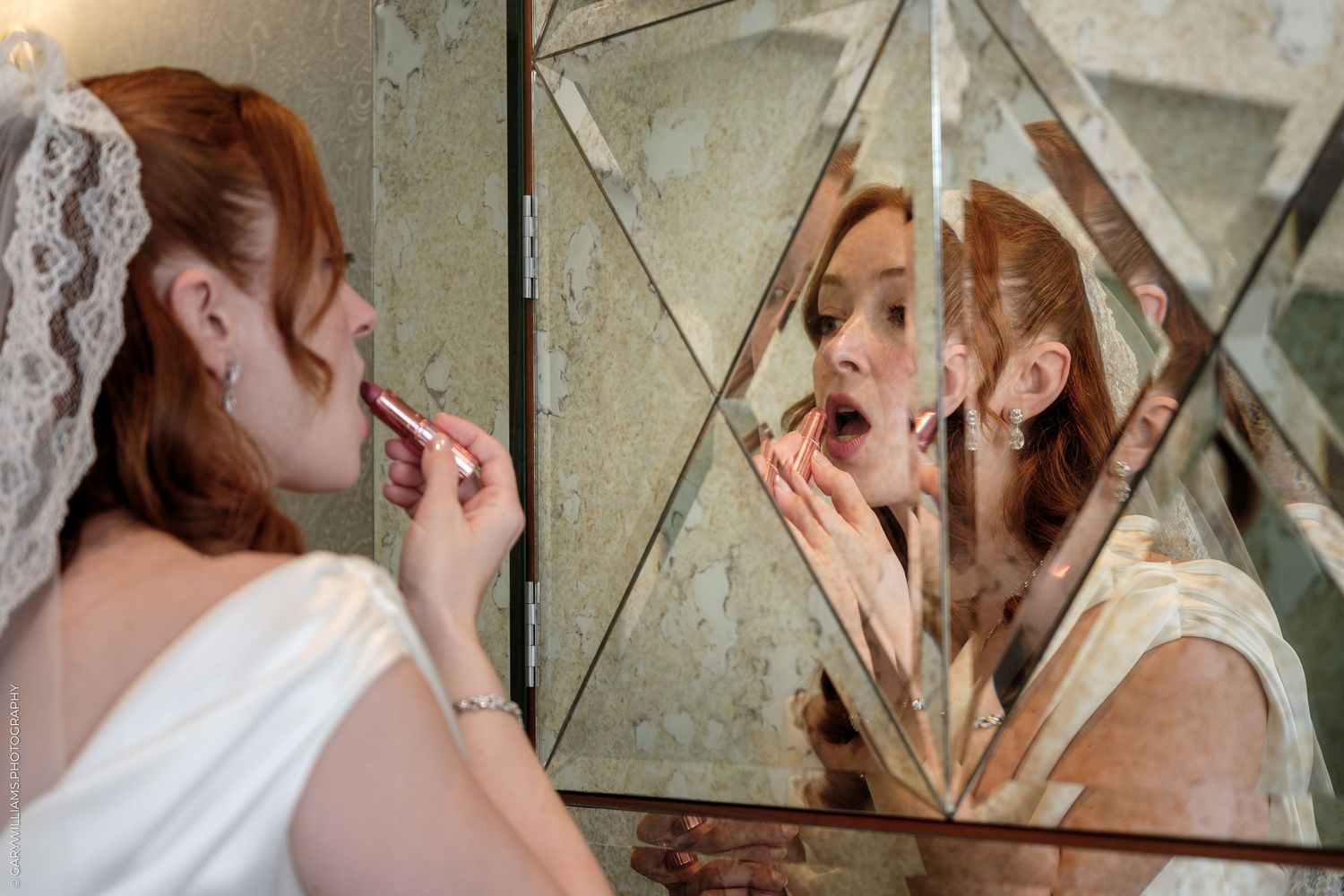 Bride applying lipstick while looking in a decorative geometric mirror, wearing a white dress and veil.