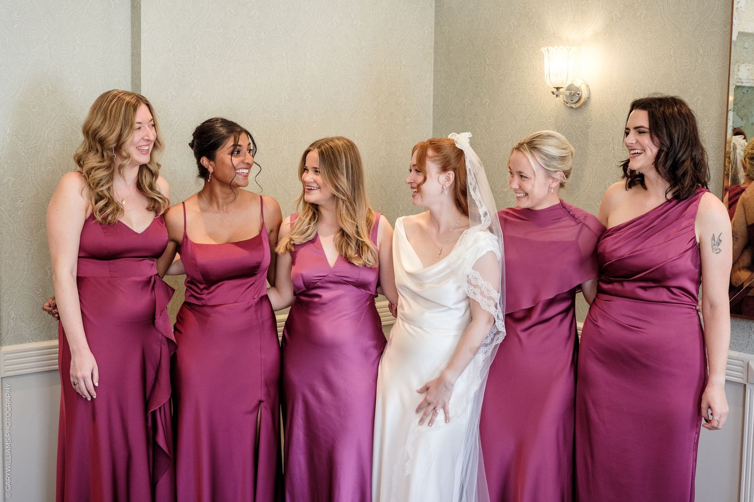 Bride in white gown laughing with five bridesmaids in mauve satin dresses at a wedding celebration.