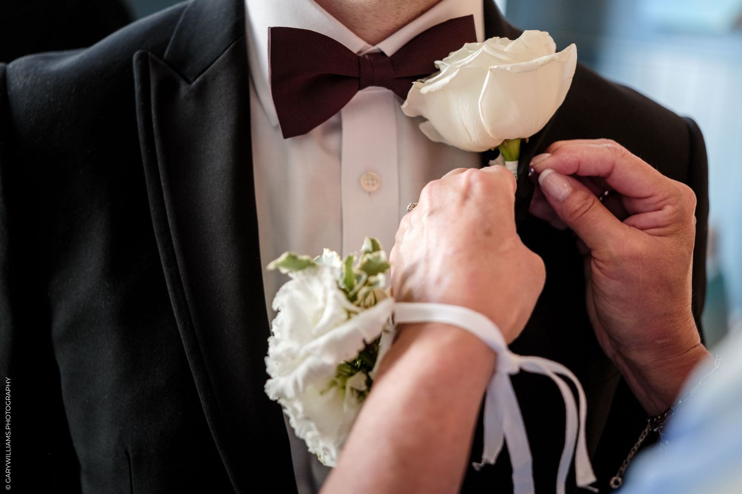 A white rose boutonniere being pinned onto a groom's black tuxedo lapel at a wedding ceremony.
