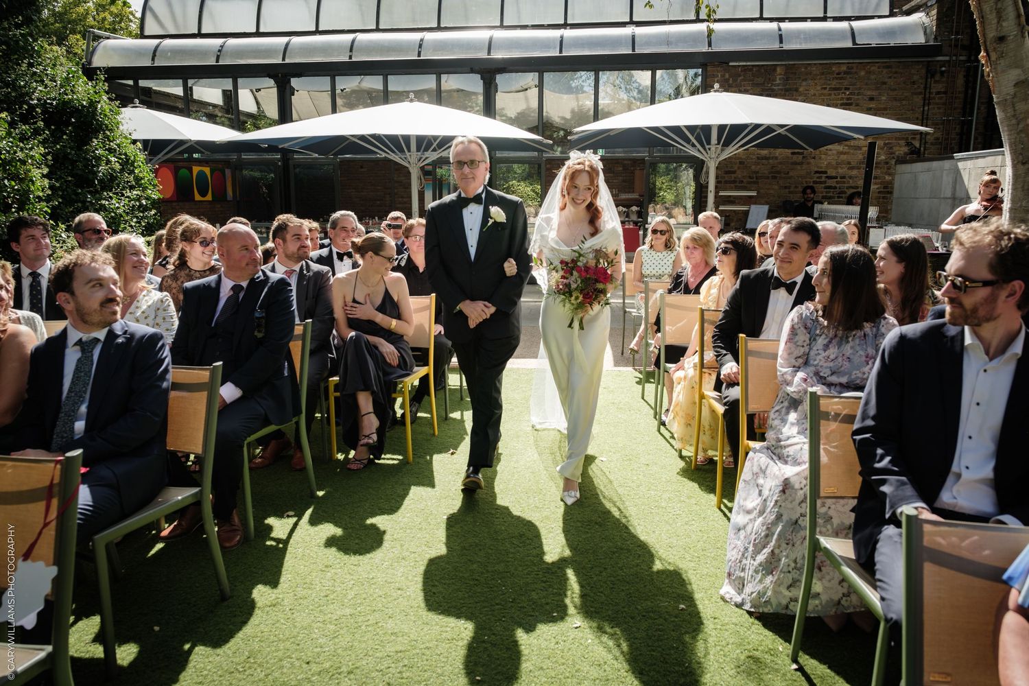 Bride walks down outdoor aisle with father at sunny wedding ceremony, guests seated on yellow chairs.