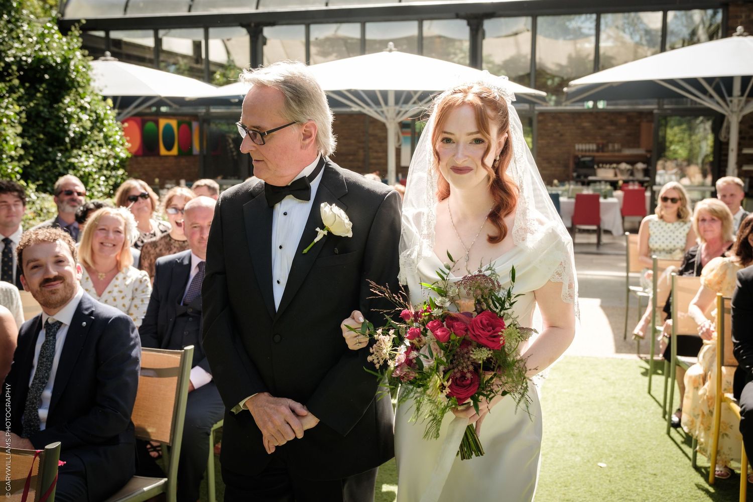 Bride in white dress and veil walks down aisle with man in tuxedo at outdoor wedding ceremony.
