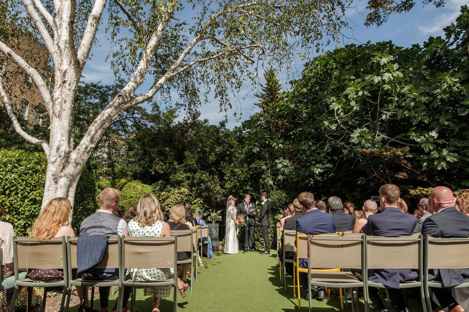 Outdoor wedding ceremony with guests seated on chairs in a lush garden beneath a white birch tree on a sunny day.