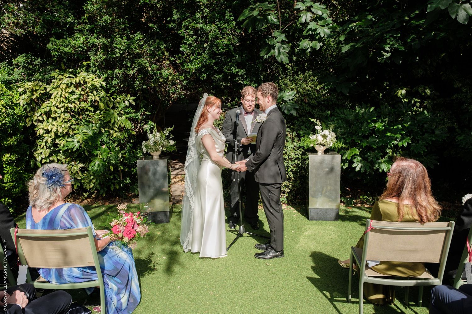 Outdoor wedding ceremony on green turf with bride and groom exchanging vows before officiant, surrounded by lush garden greenery.