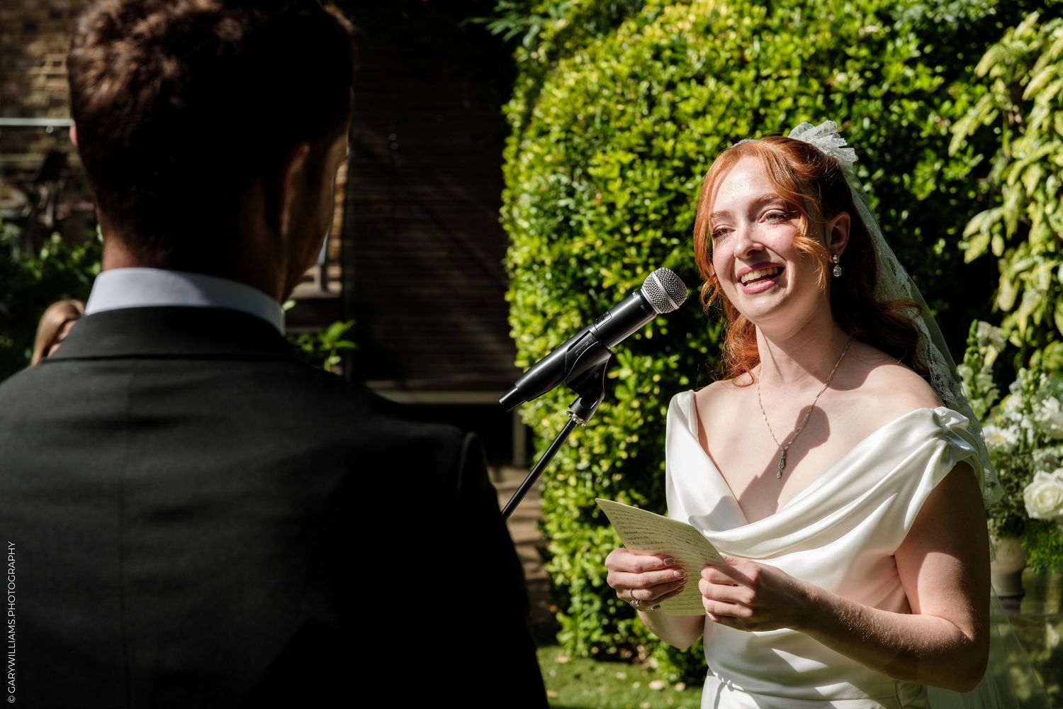 Smiling bride in white gown reads vows at outdoor wedding ceremony with lush green garden backdrop.
