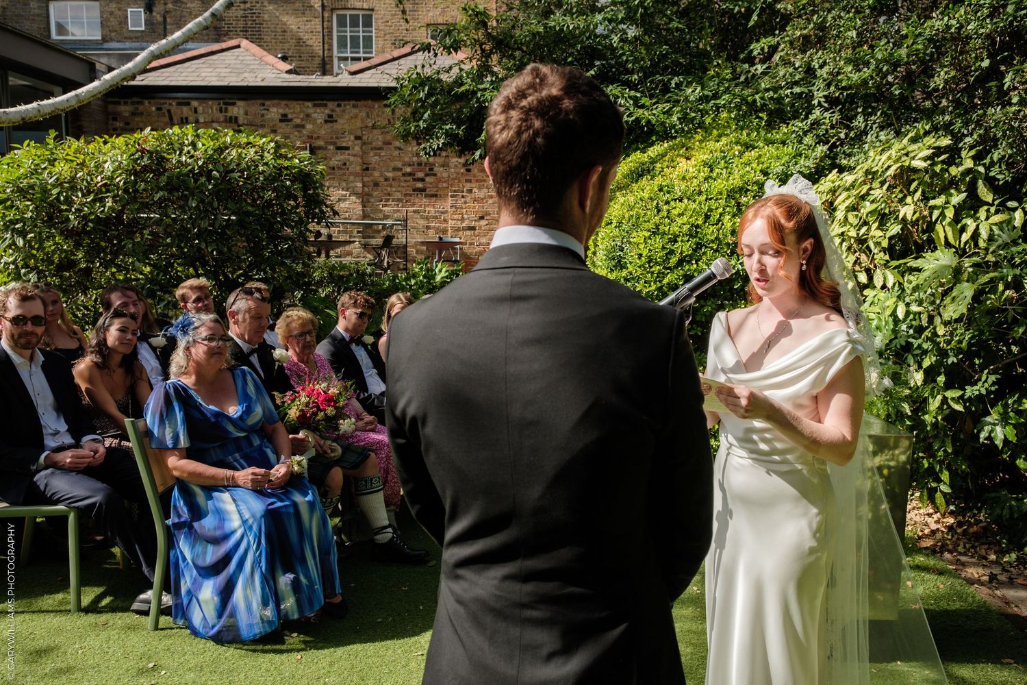 Bride reads vows to groom during outdoor wedding ceremony with seated guests in garden setting.