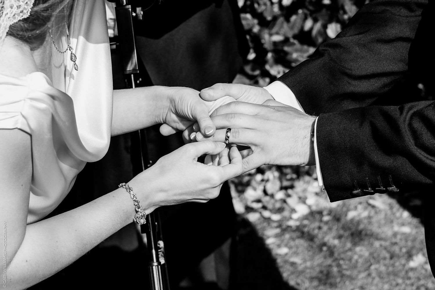 Black and white photo of bride placing wedding ring on groom's finger during outdoor ceremony.