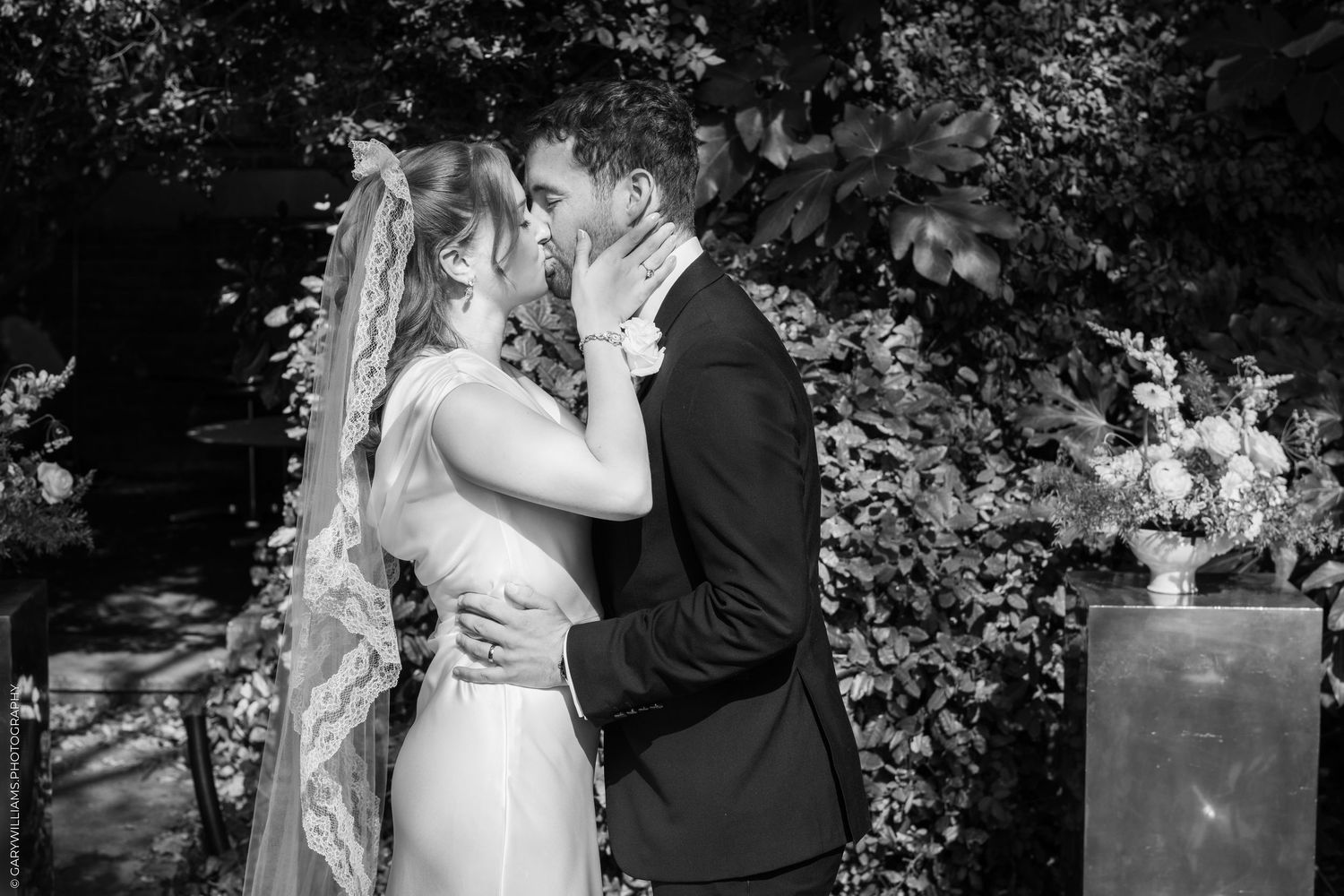 Black and white photo of a bride and groom sharing a first kiss at their outdoor wedding ceremony.