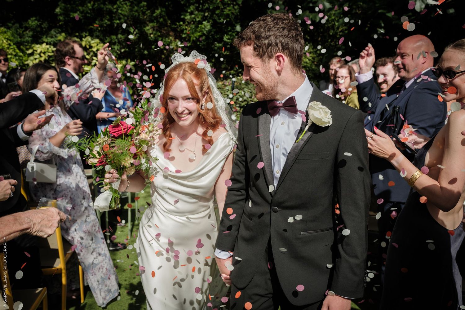 Bride and groom walk through confetti shower after outdoor wedding ceremony, bride holding colorful bouquet.