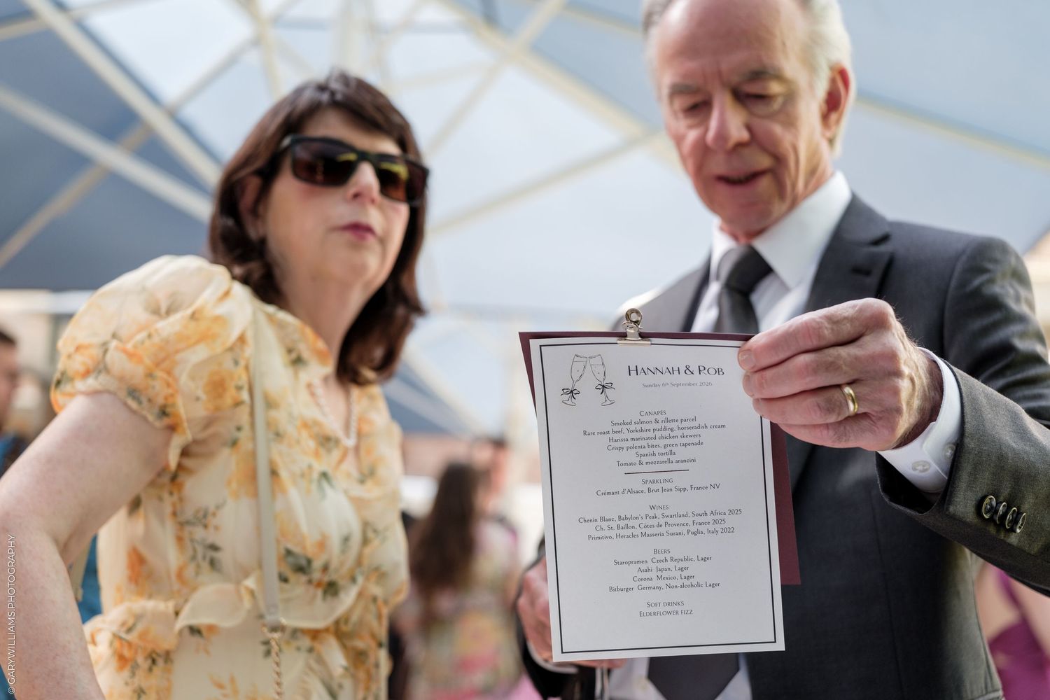 Man and woman examining a framed certificate at an outdoor ceremony, woman in floral dress stands beside him.