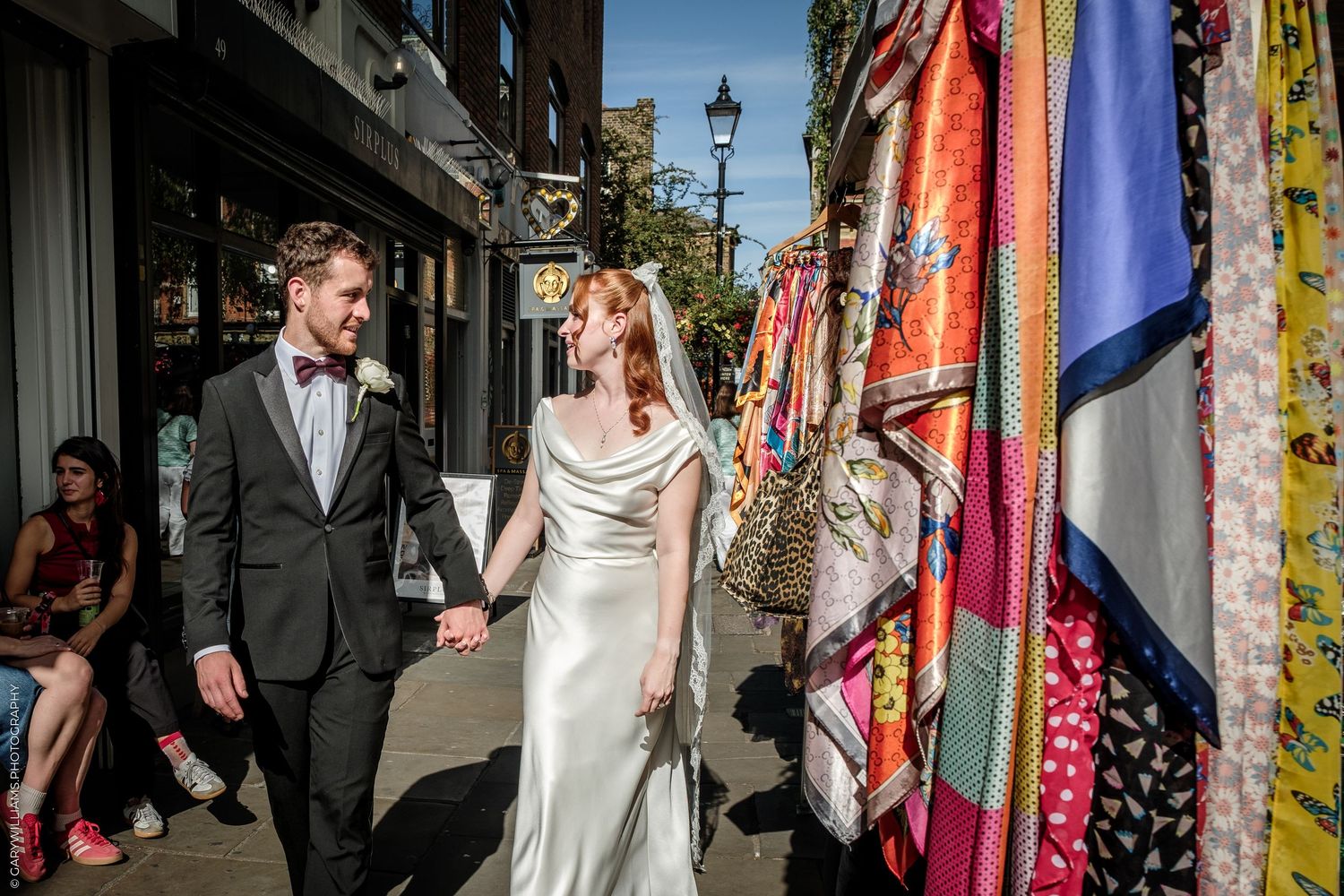 A bride and groom hold hands while walking past colorful fabric displays on a sunny cobblestone street.