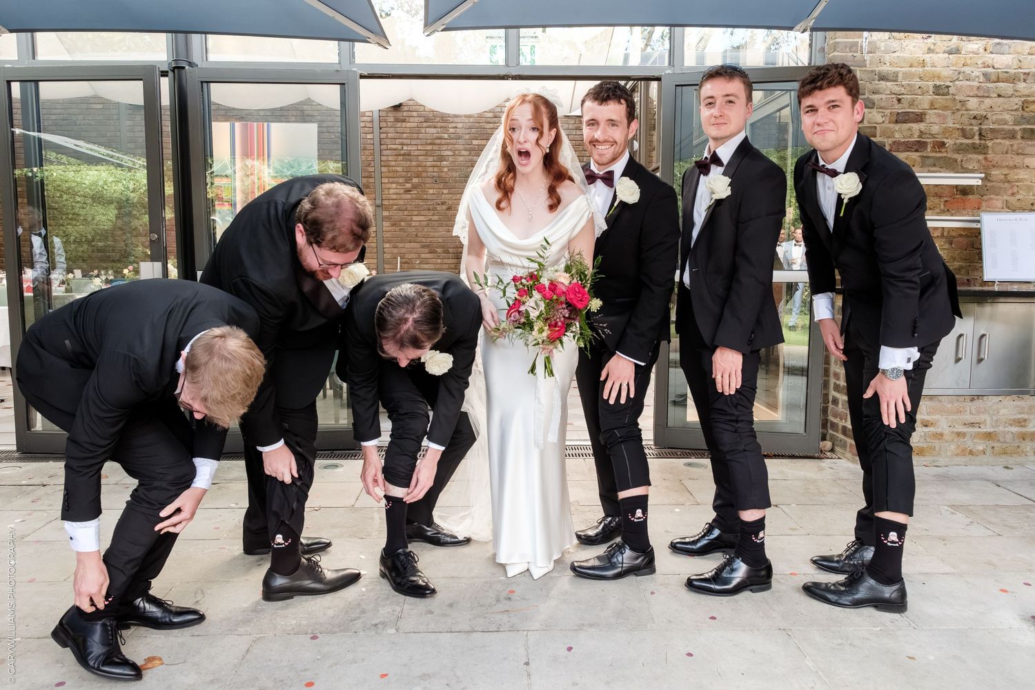 Bride and groom pose with groomsmen in black tuxedos, some bending down, at an outdoor wedding venue.