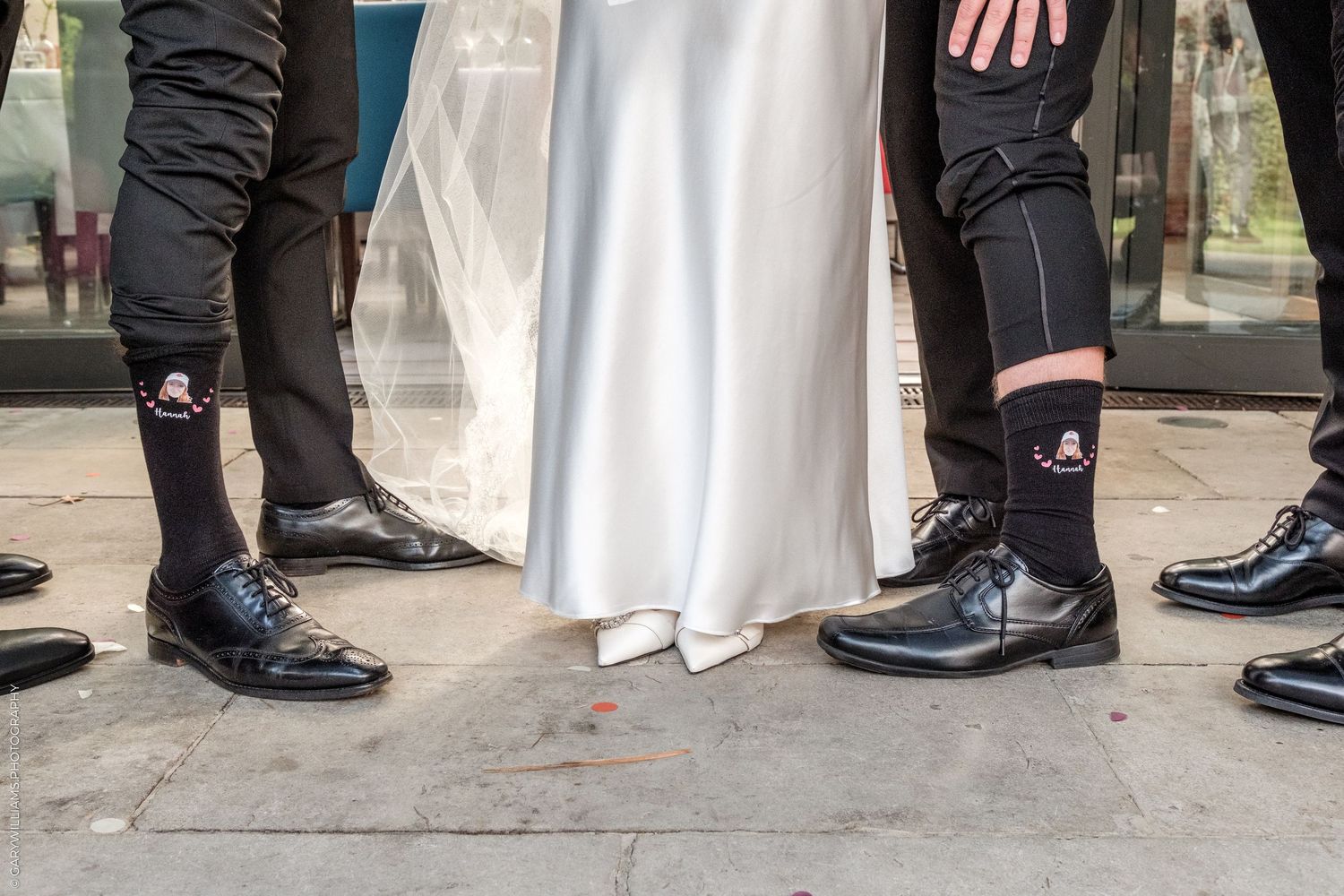 Close-up of wedding party feet showing black dress shoes and custom black socks with logo, beside a bride in white gown.
