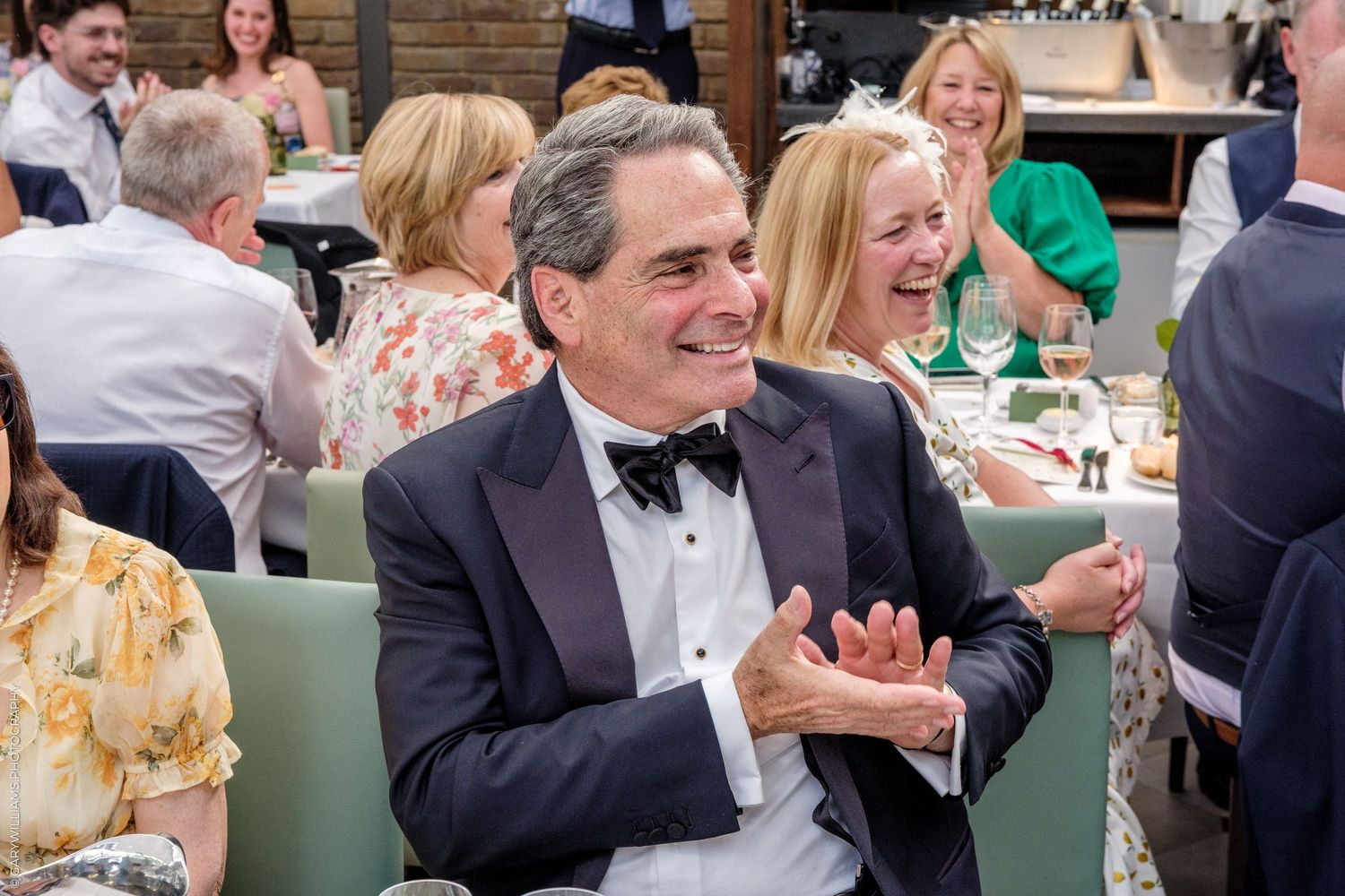 Man in tuxedo smiling and clapping at elegant dinner event surrounded by guests in formal attire.