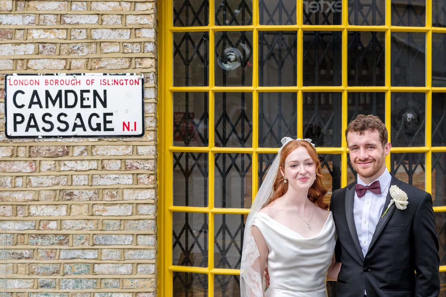 Bride and groom pose by Camden Passage N1 street sign in London, bride in white gown, groom in black tuxedo.