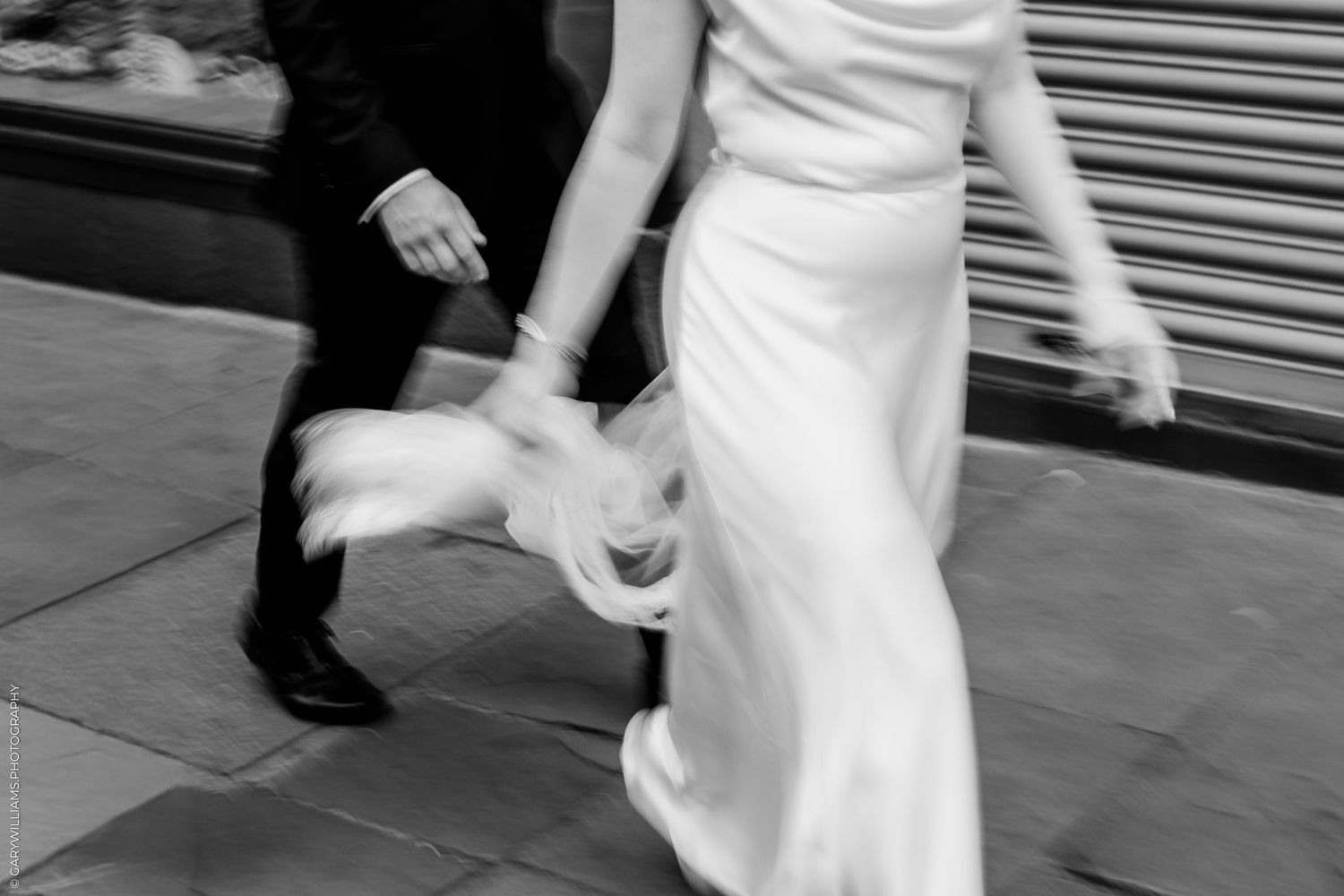 Black and white photo of a bride and groom walking briskly on a city sidewalk, dress flowing with motion blur.