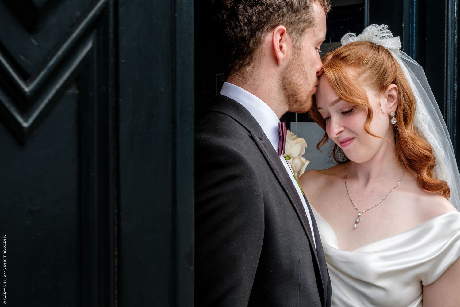 Bride and groom share an intimate moment by a dark door, bride in white gown with veil, groom in gray suit.