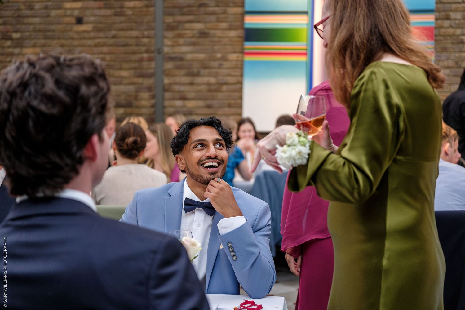 A smiling man in a light blue suit and bow tie chats with guests at an elegant indoor wedding reception.