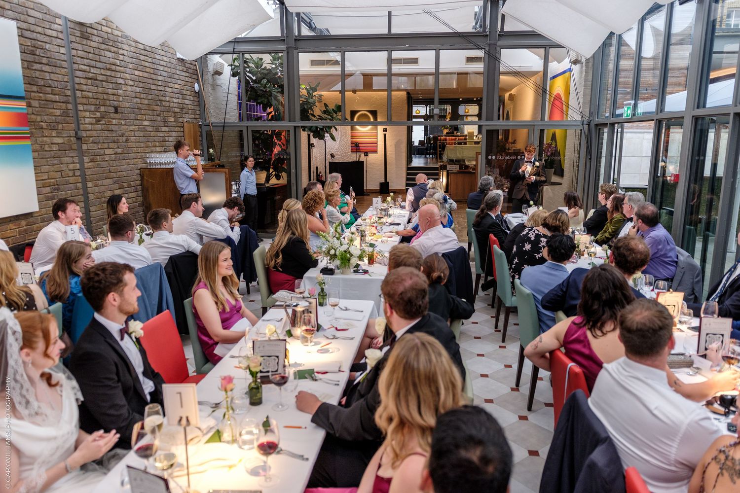 Elegant indoor wedding reception dinner with guests seated at long decorated tables in a bright venue.