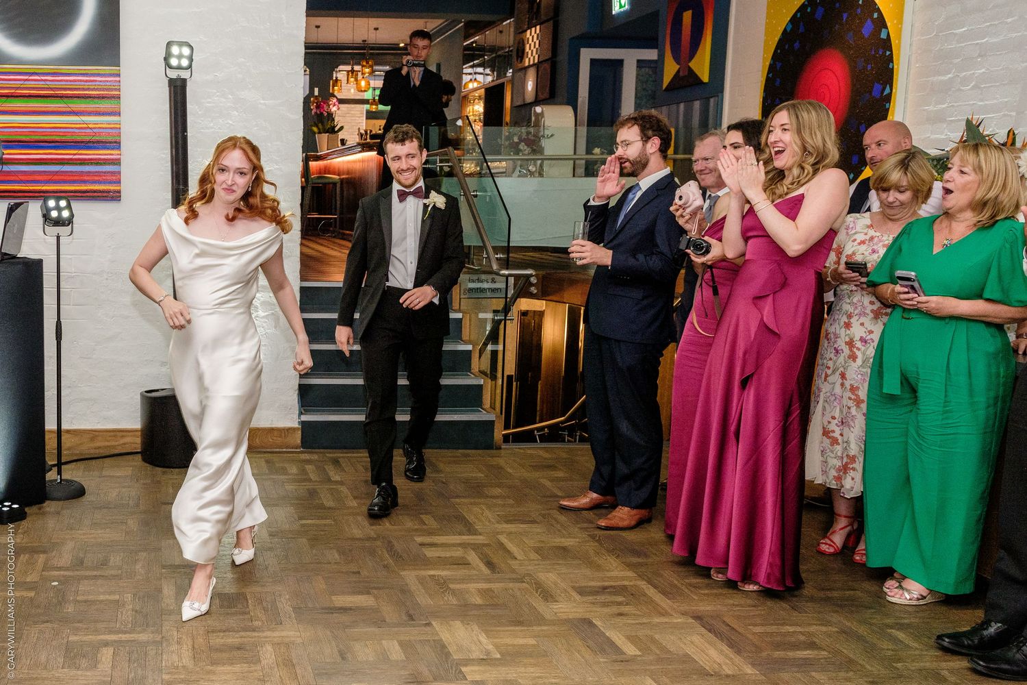 Bride in white jumpsuit and groom in tuxedo make grand entrance as wedding guests cheer and applaud in elegant venue.