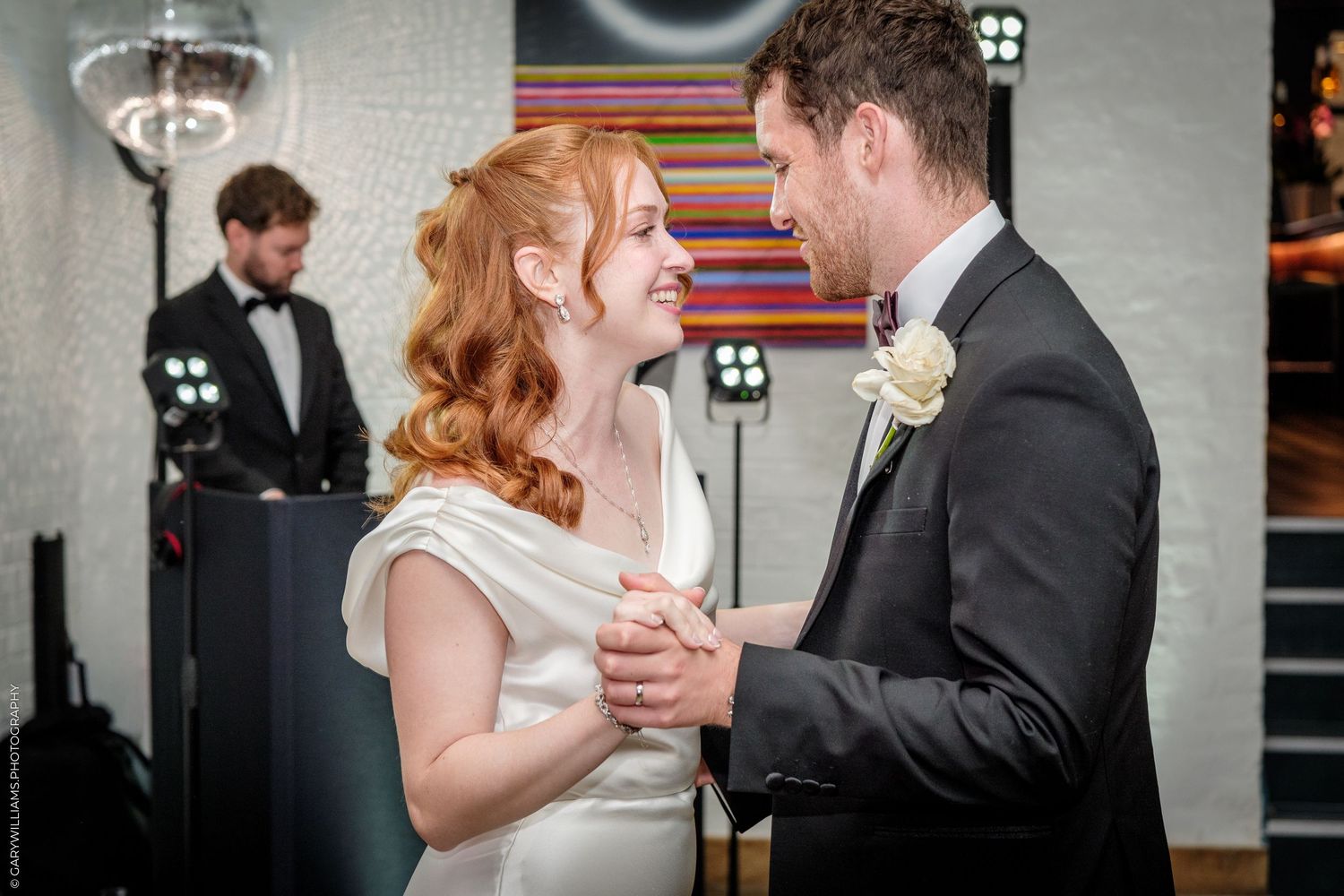 Bride and groom share their first dance, smiling joyfully, with a DJ in the background at their wedding reception.