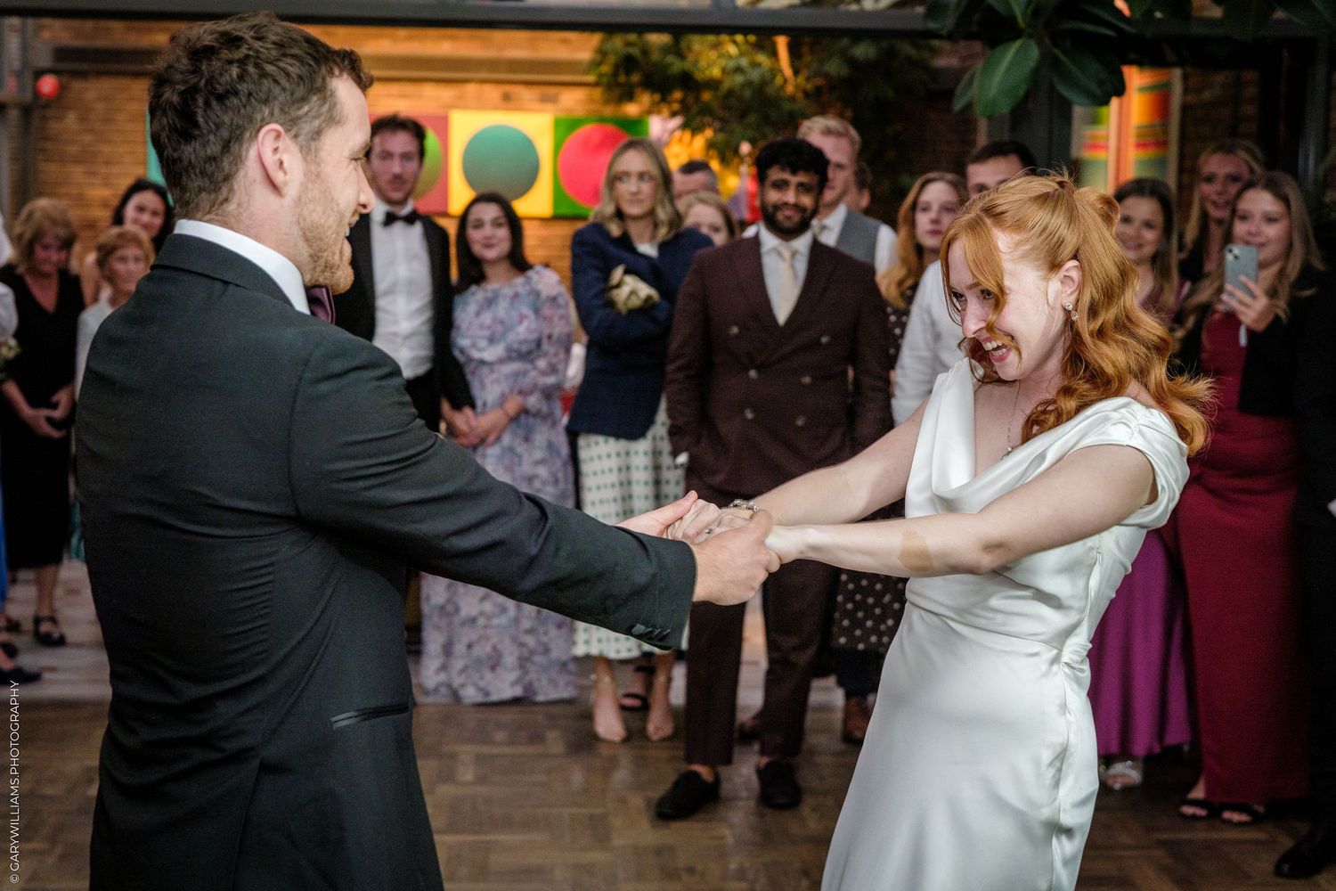 Bride and groom share first dance holding hands, surrounded by smiling wedding guests in elegant venue.