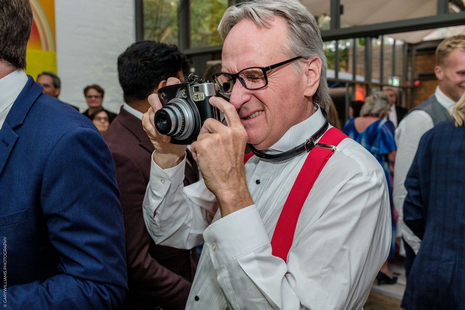 Smiling man in white shirt with red suspenders holding a film camera at a formal indoor event.