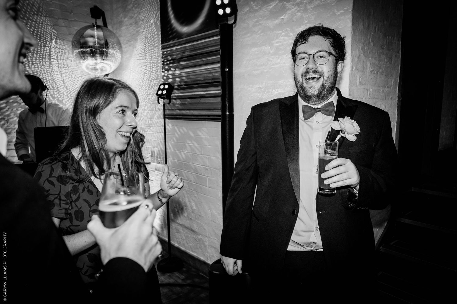 Black and white photo of a man in a tuxedo and woman laughing at a wedding reception holding drinks.