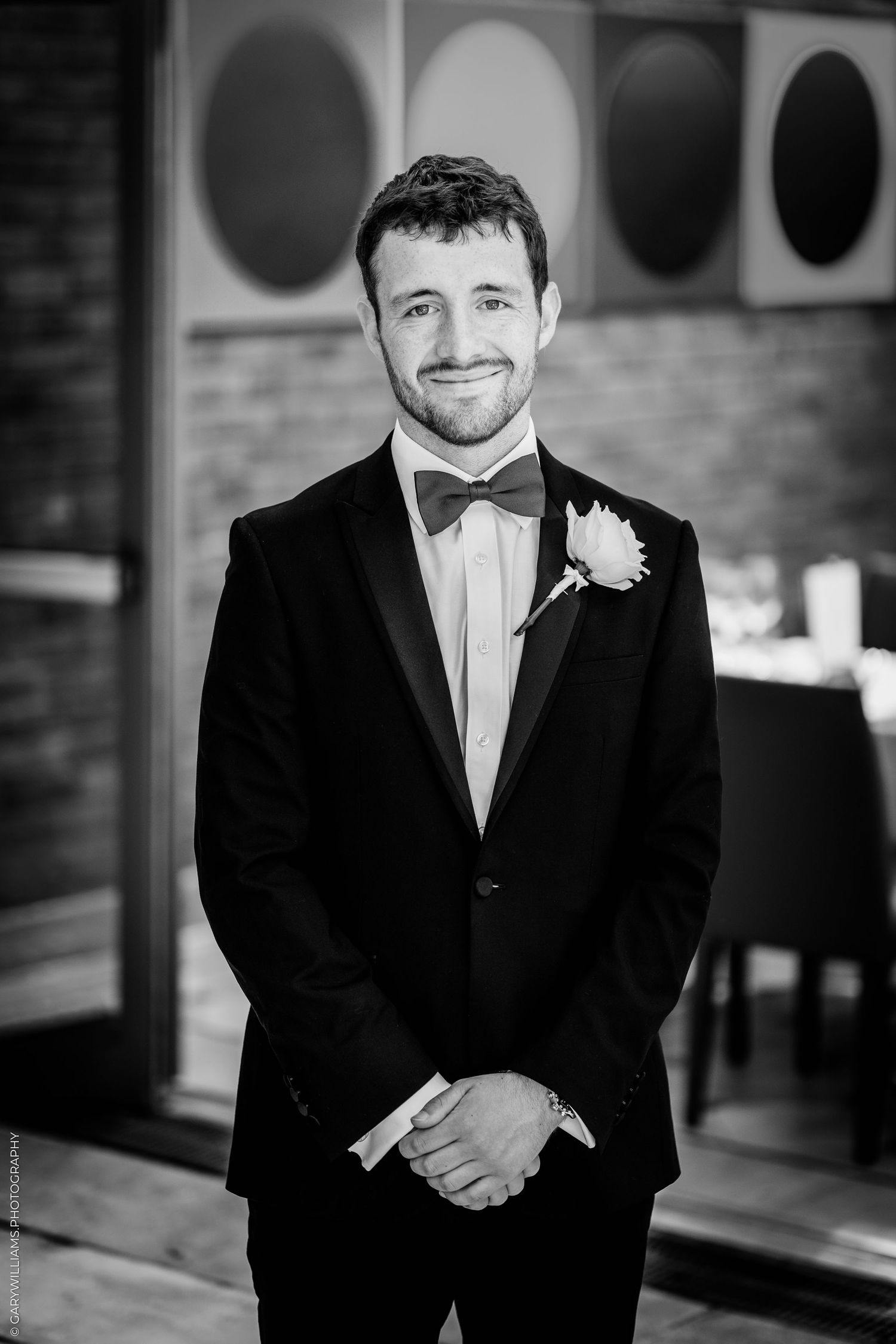 A smiling groom in a black tuxedo with bow tie and boutonniere poses for a black and white portrait photo.