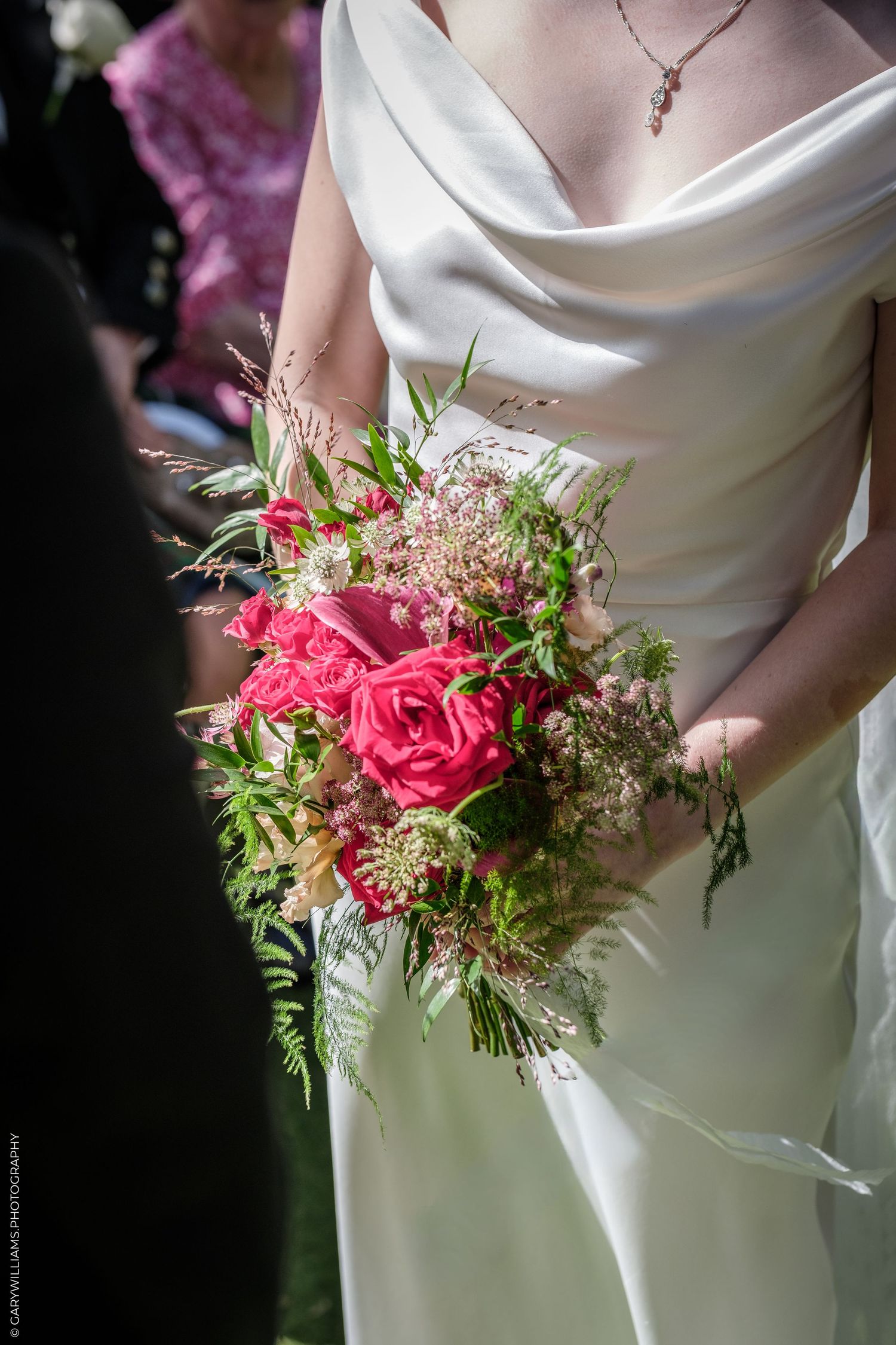 Bride in white gown holding a lush pink rose and wildflower bouquet with ferns at a wedding ceremony.