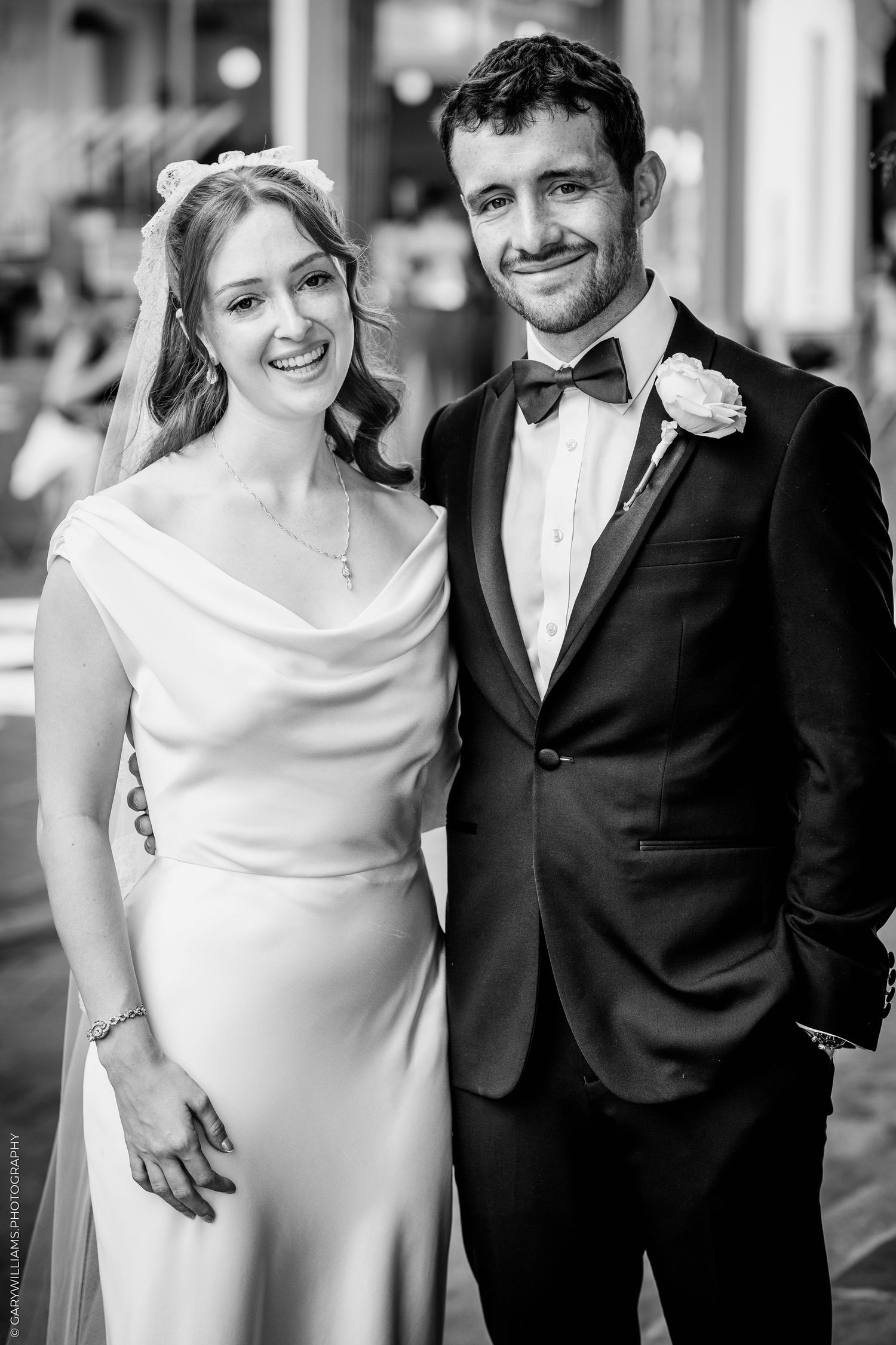 Black and white photo of a smiling bride in white gown and groom in tuxedo posing together at their wedding reception.