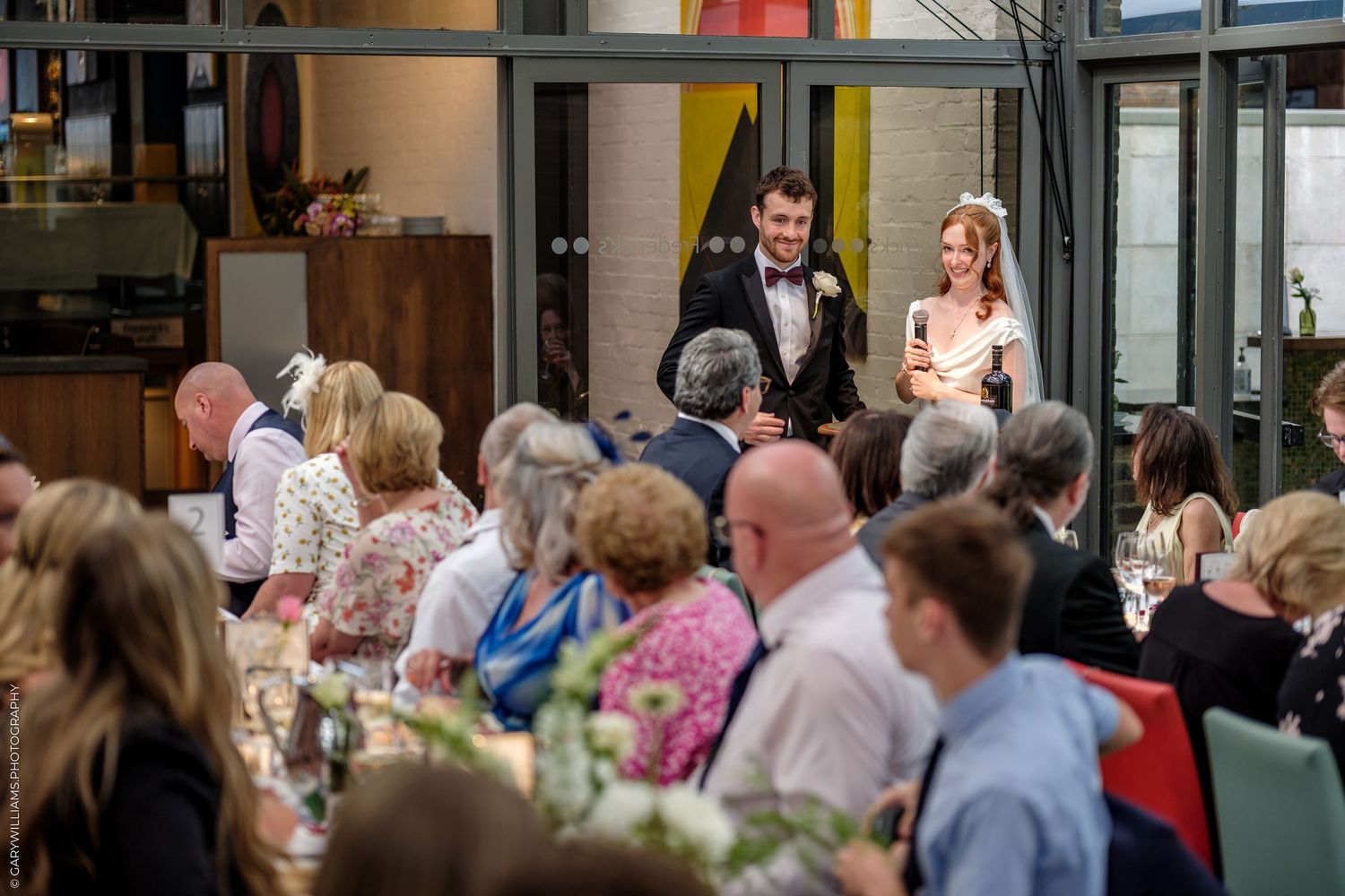 Bride and groom stand smiling before wedding reception guests seated at decorated tables in a modern venue.
