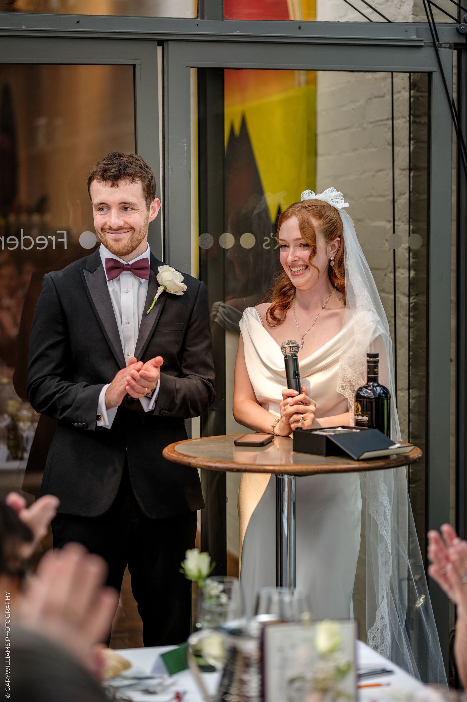 Smiling bride holding microphone at wedding reception podium beside groom in black tuxedo with bow tie.