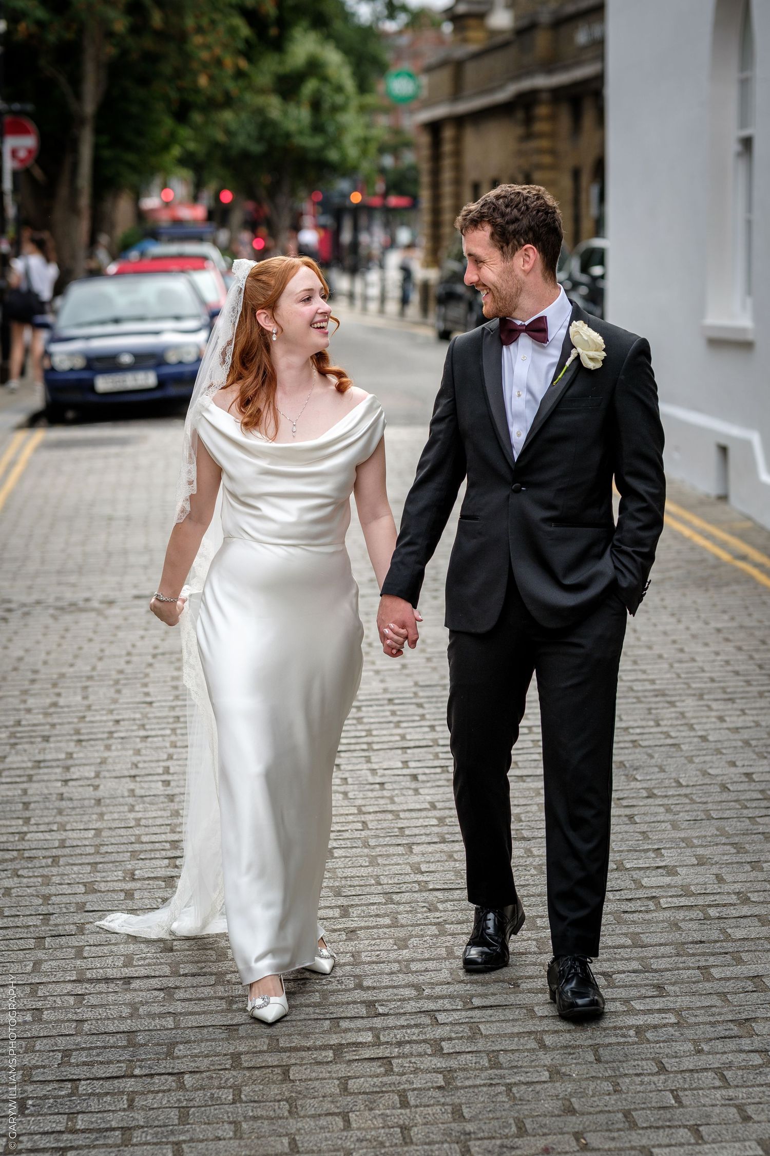 A bride and groom walk hand-in-hand on a cobblestone street, smiling at each other in wedding attire.