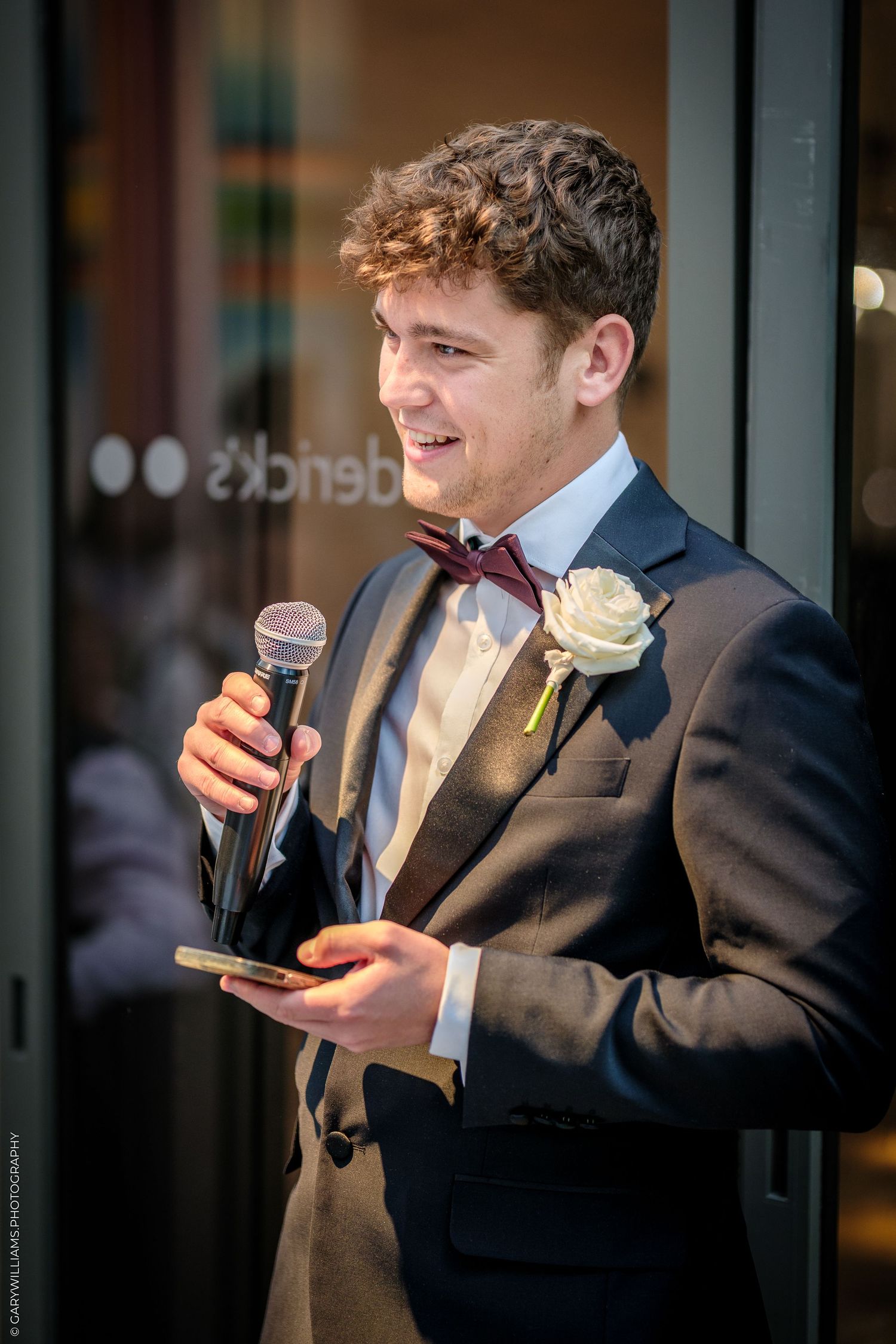 Young man in a dark suit with bow tie and boutonniere holds a microphone while giving a speech at a wedding.