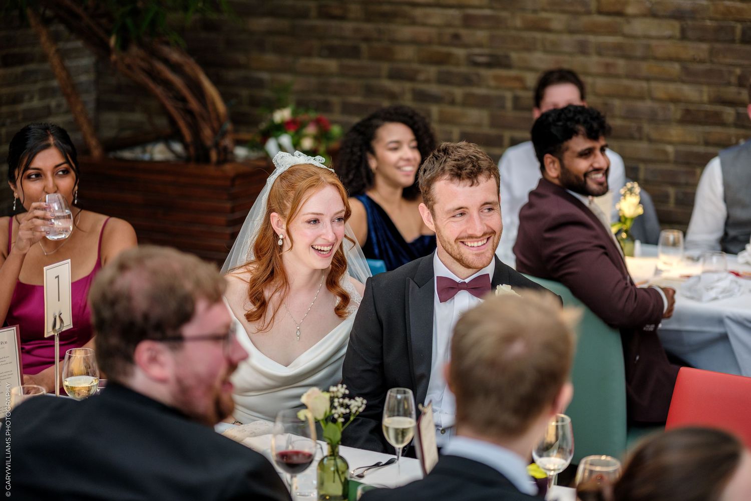 Smiling bride and groom sit together at wedding reception table surrounded by guests in elegant venue.