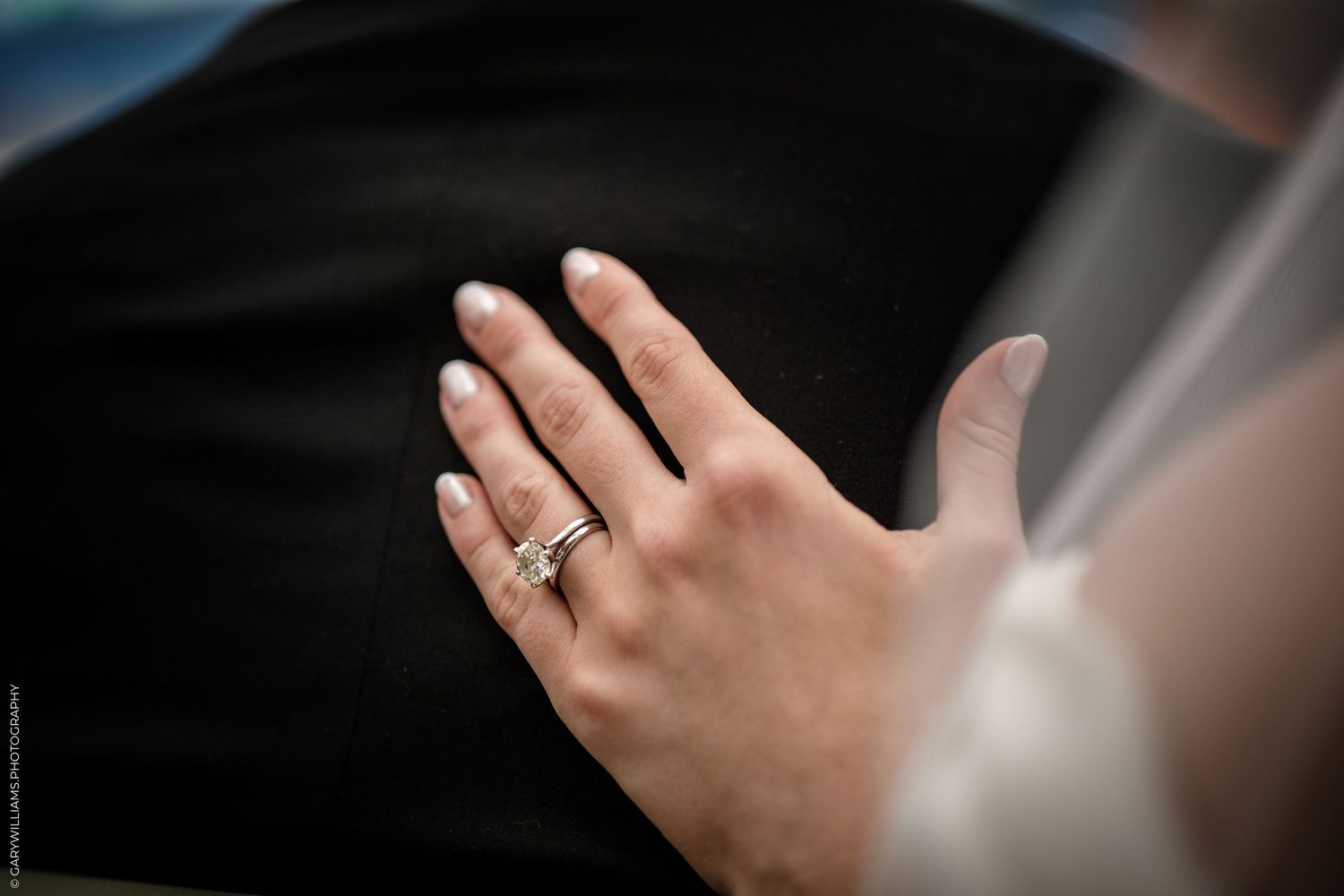 Close-up of a bride's hand displaying a diamond engagement ring and wedding band against a dark suit jacket.