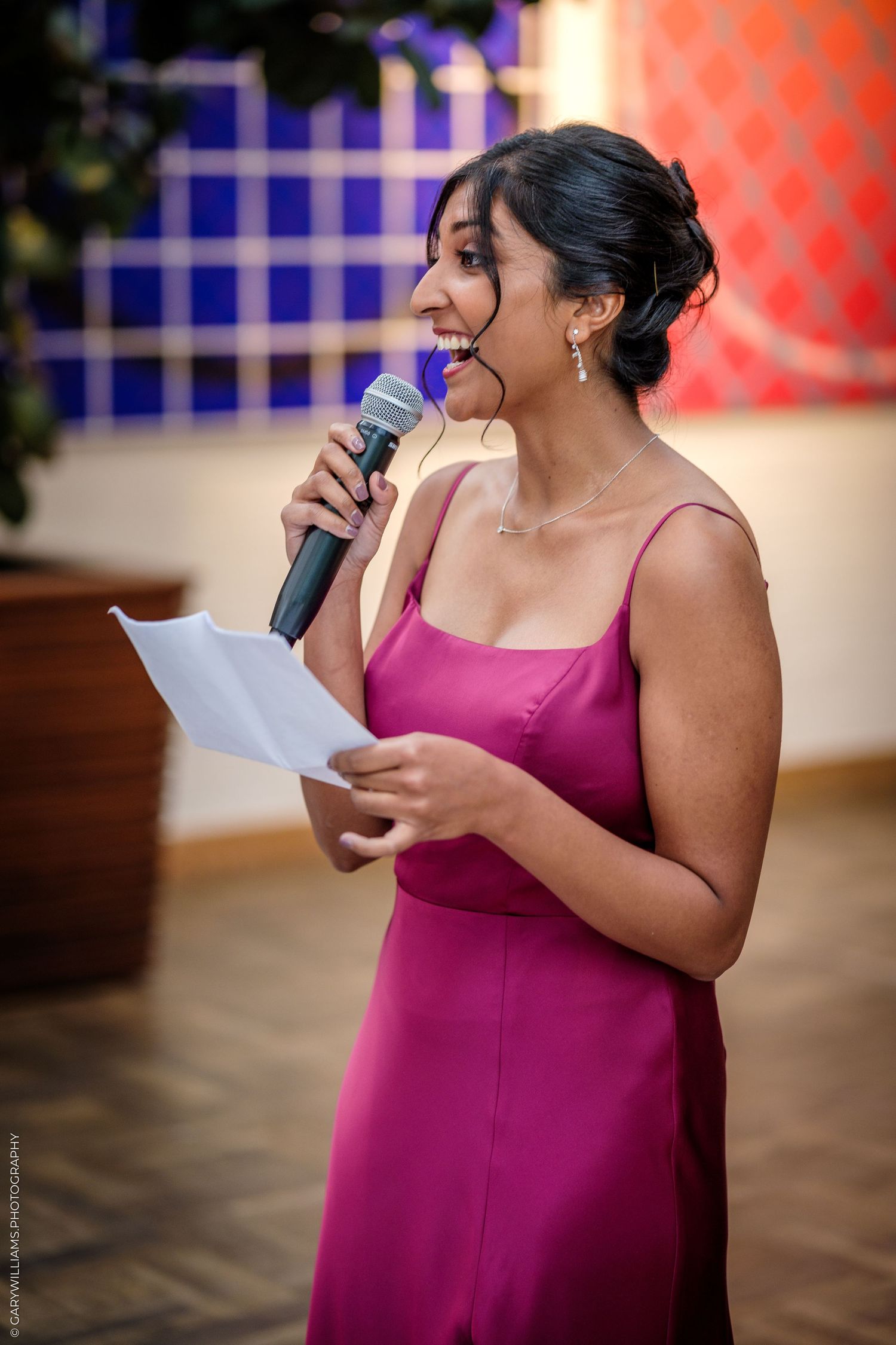 Woman in pink dress giving speech at event, holding microphone and notes, colorful art backdrop.