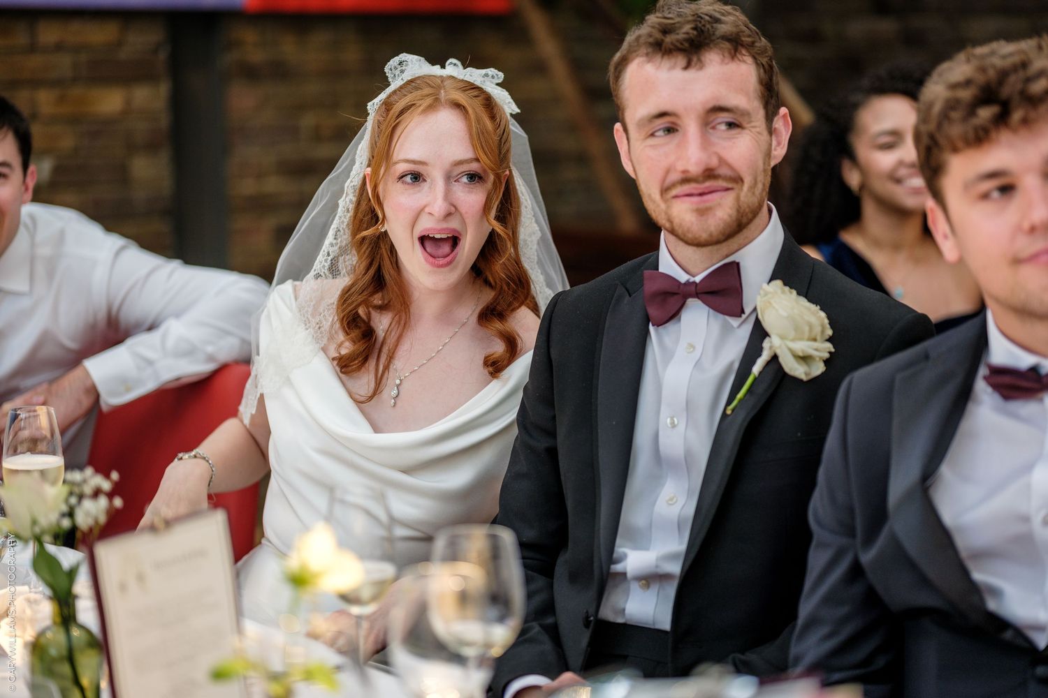 Bride and groom smiling at wedding reception table, bride wearing veil, groom in dark suit with boutonniere.