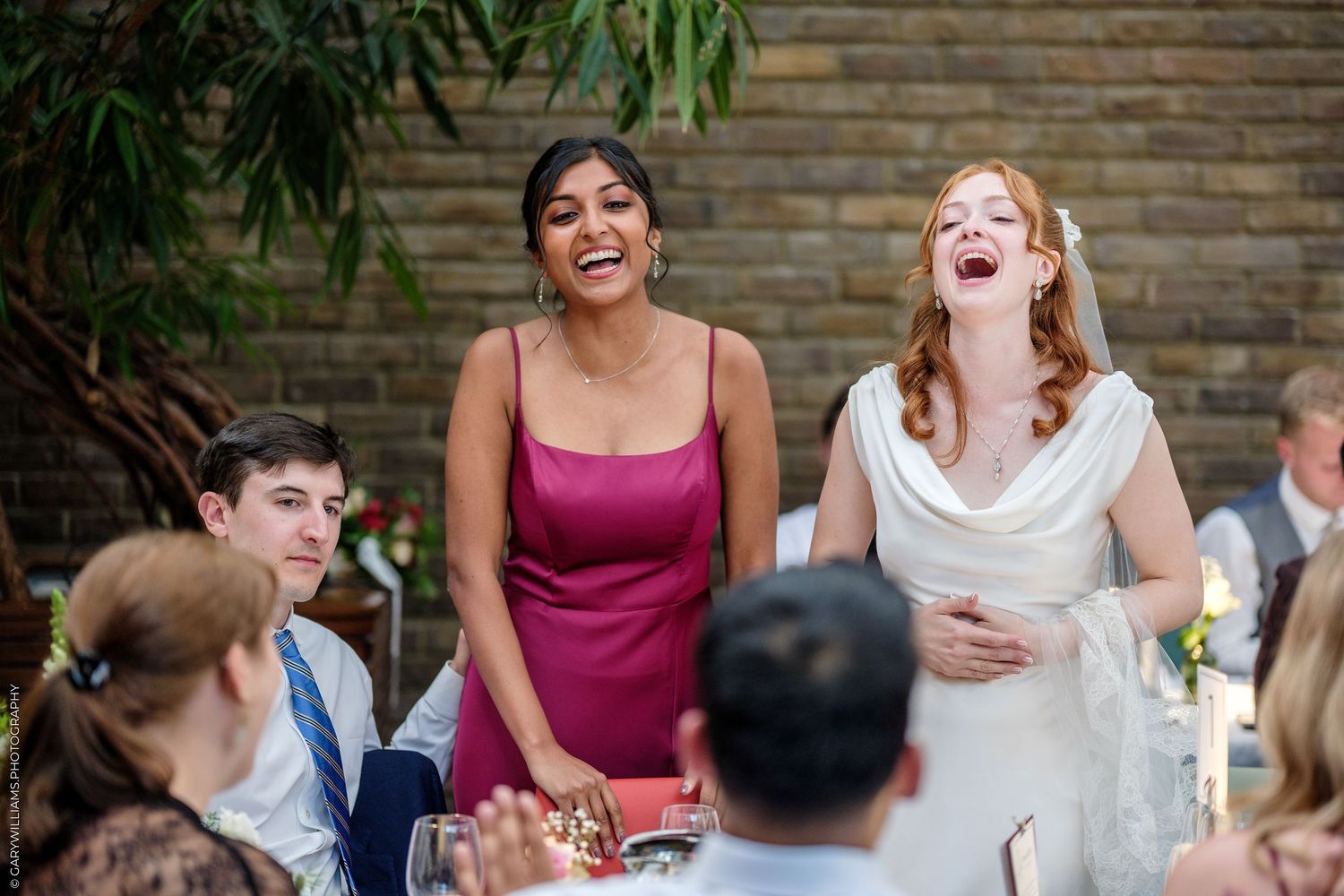 Bride and bridesmaid laughing joyfully at wedding reception table, wearing white gown and magenta dress.
