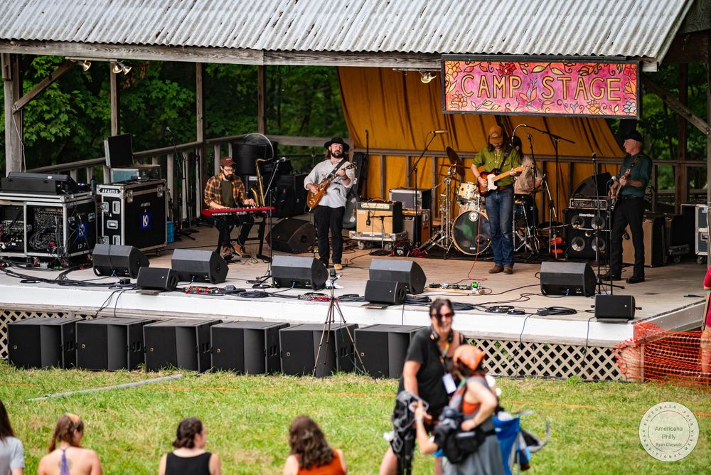 Live band performing on outdoor stage at a sunny music festival with audience gathered on green lawn.